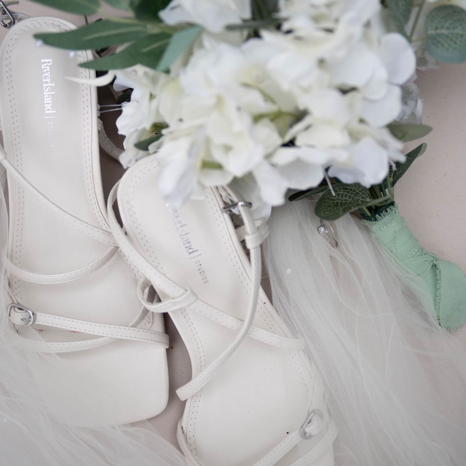 White strappy bridal heels beside a bouquet of white hydrangeas and greenery on a soft white surface.