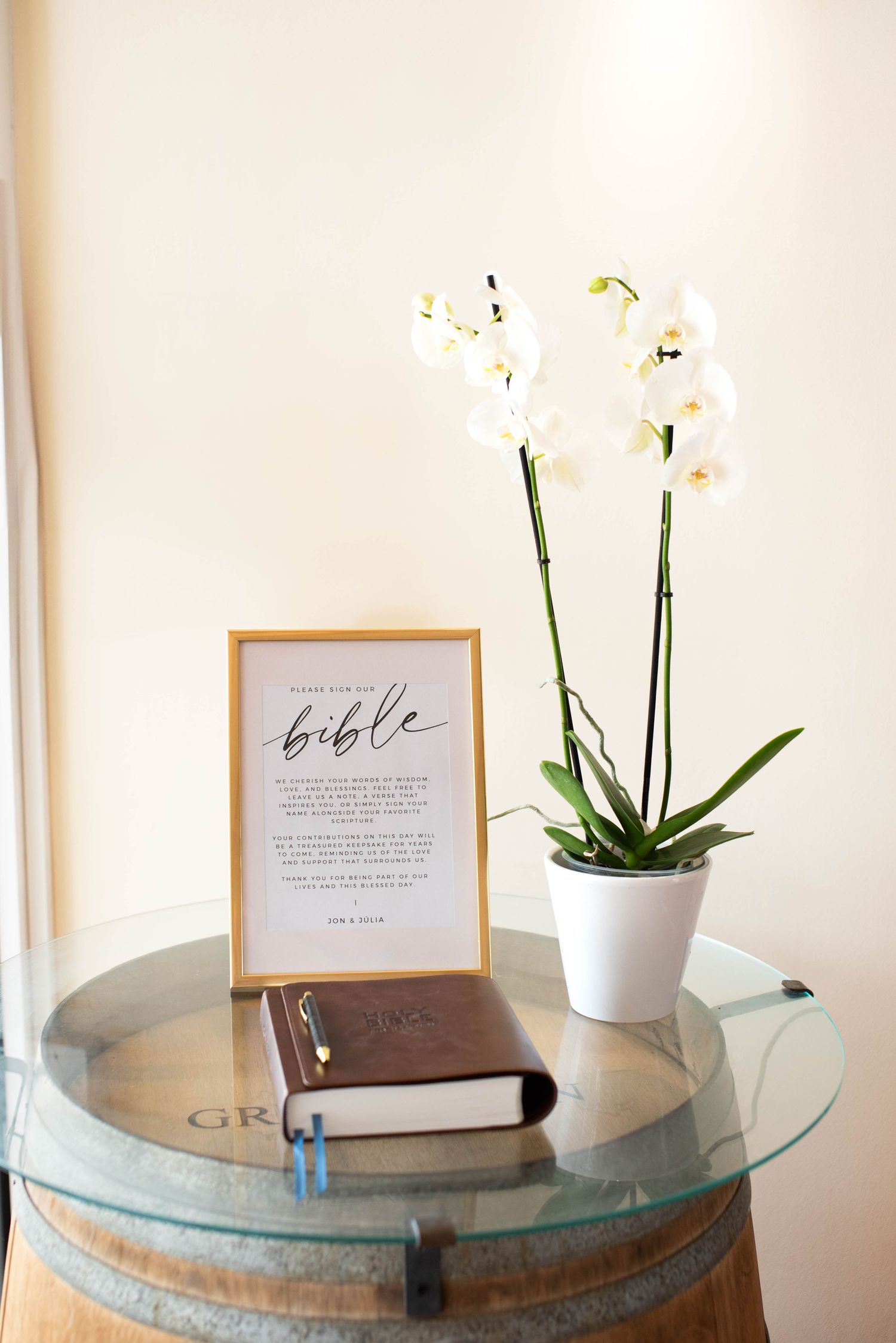 Wedding bible guest book display with white orchid plant and framed sign on barrel table.