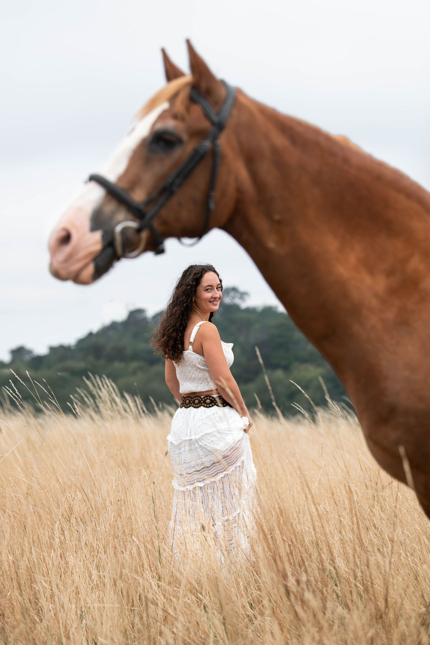 Woman in white dress smiling beside a chestnut horse in a golden wheat field.