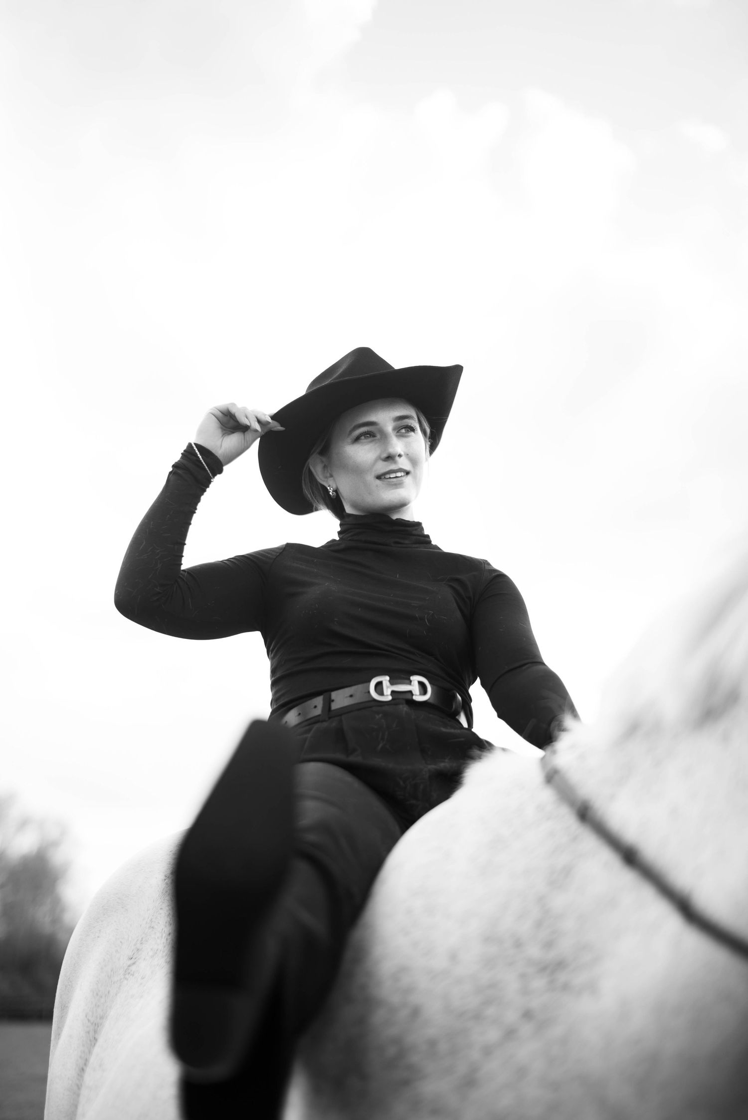 Black and white photo of a smiling woman in black riding a white horse while tipping her cowboy hat.