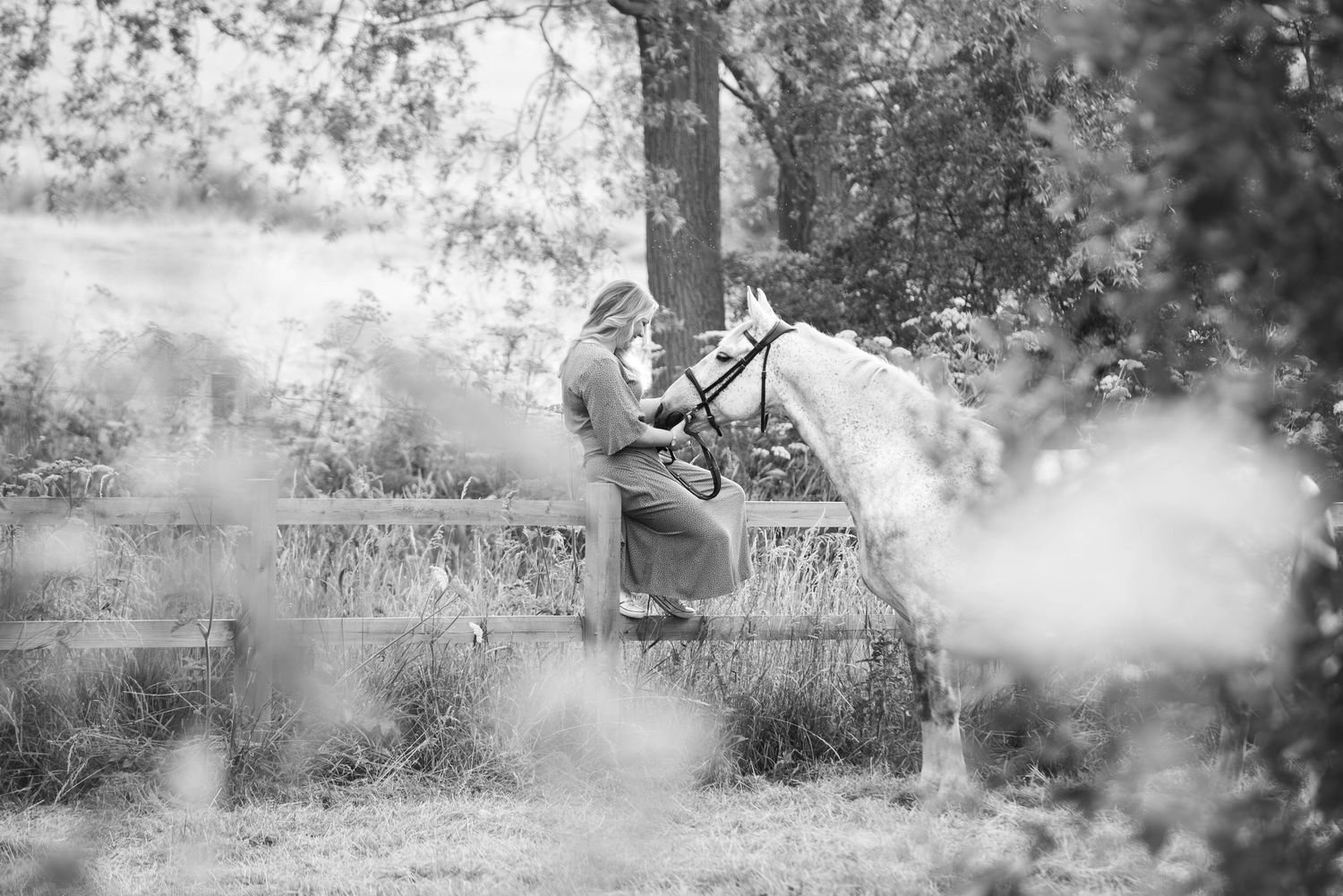 Black and white photo of woman sitting on fence with white horse in nature.