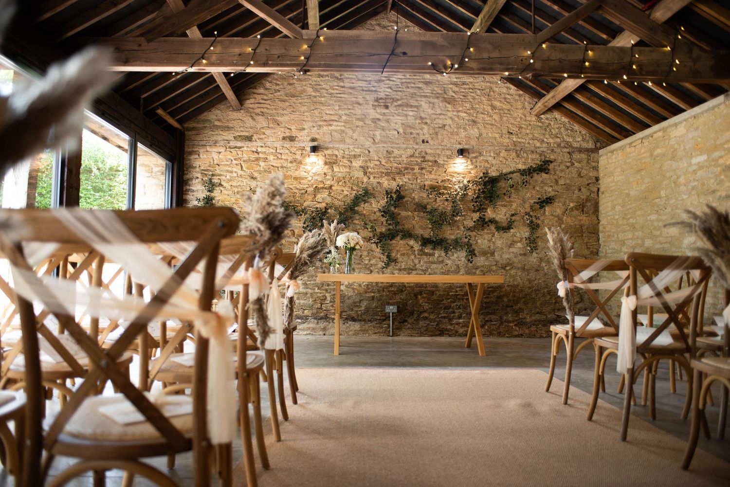 Rustic barn wedding ceremony venue with exposed stone walls, fairy lights, wooden cross-back chairs, and floral greenery backdrop.