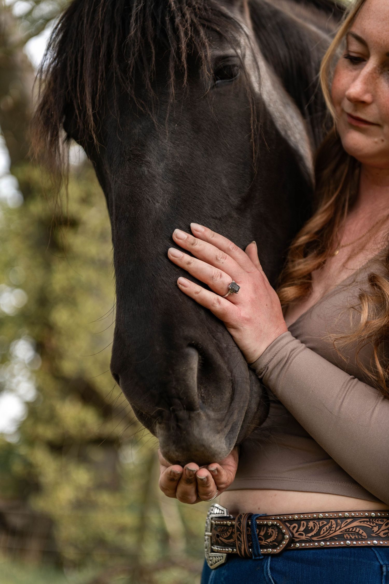Woman in western belt gently cradles dark horse's nose in her hands outdoors near green trees.