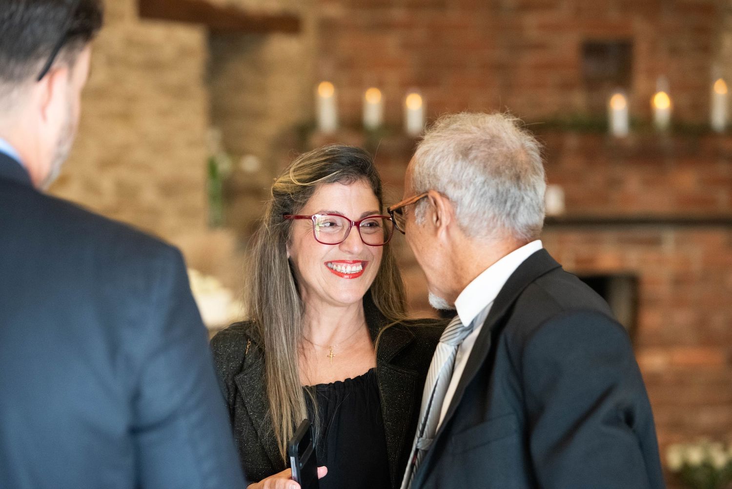 A woman in glasses smiles warmly while conversing with guests at an elegant candlelit evening event.