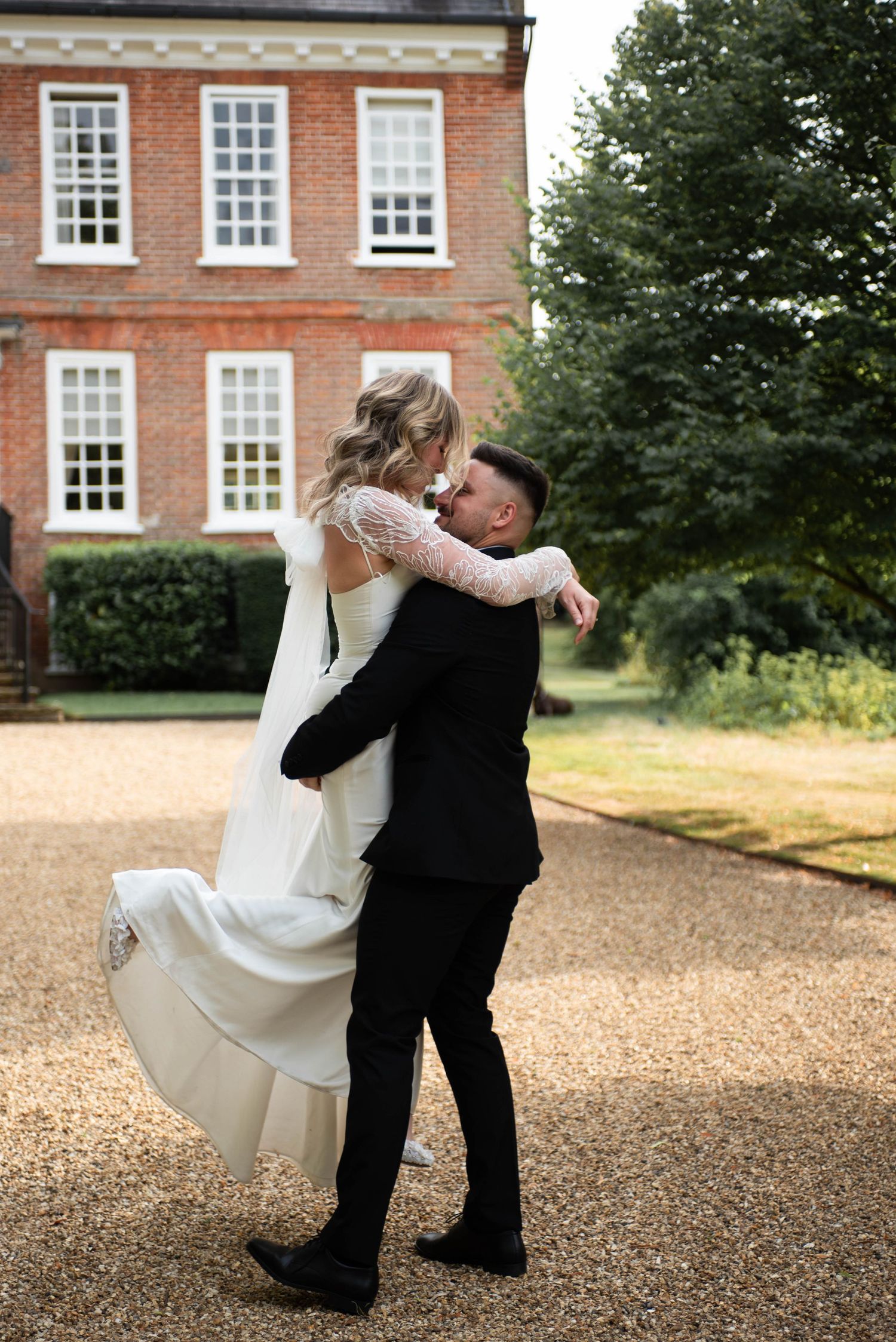 Groom lifts bride in white lace gown outside a classic red brick manor house on their wedding day.