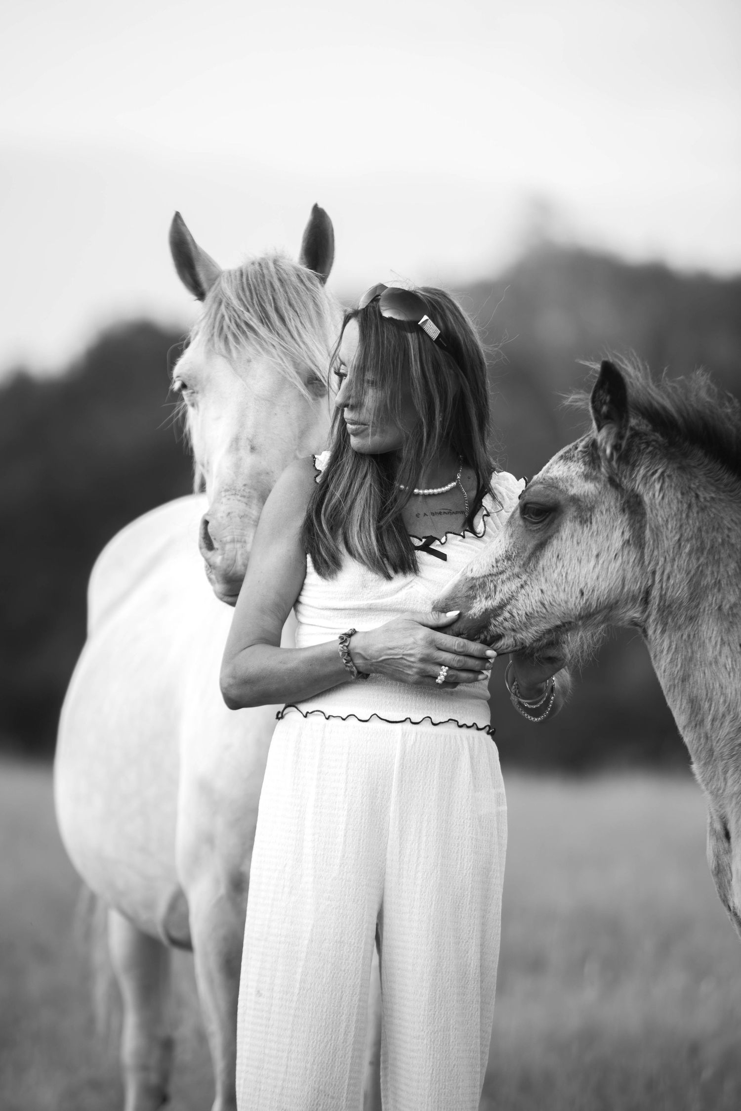 Black and white image of a woman in white standing between a white horse and a foal.