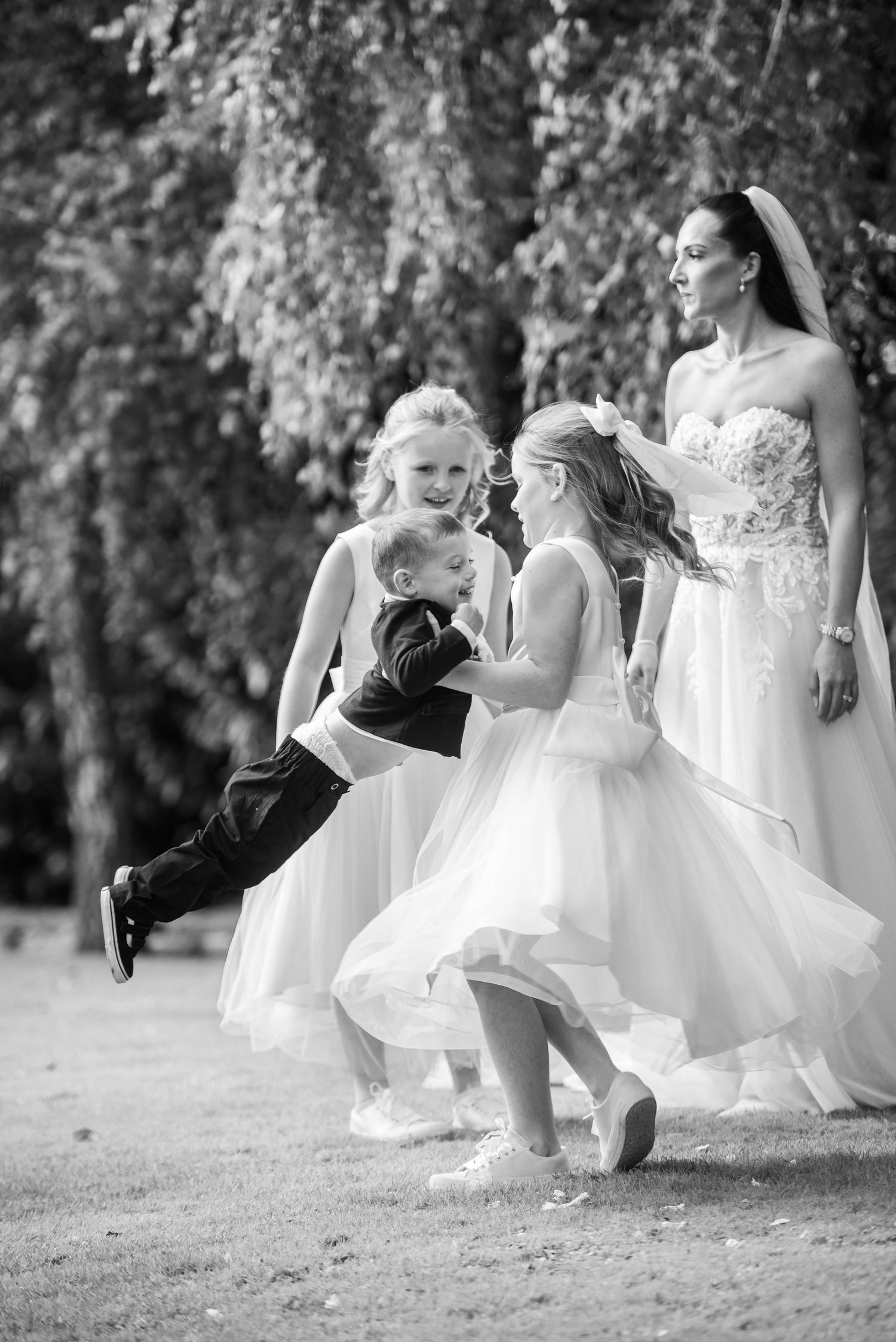 Black and white photo of a bride watching flower girls and ring bearer playing outdoors under trees.