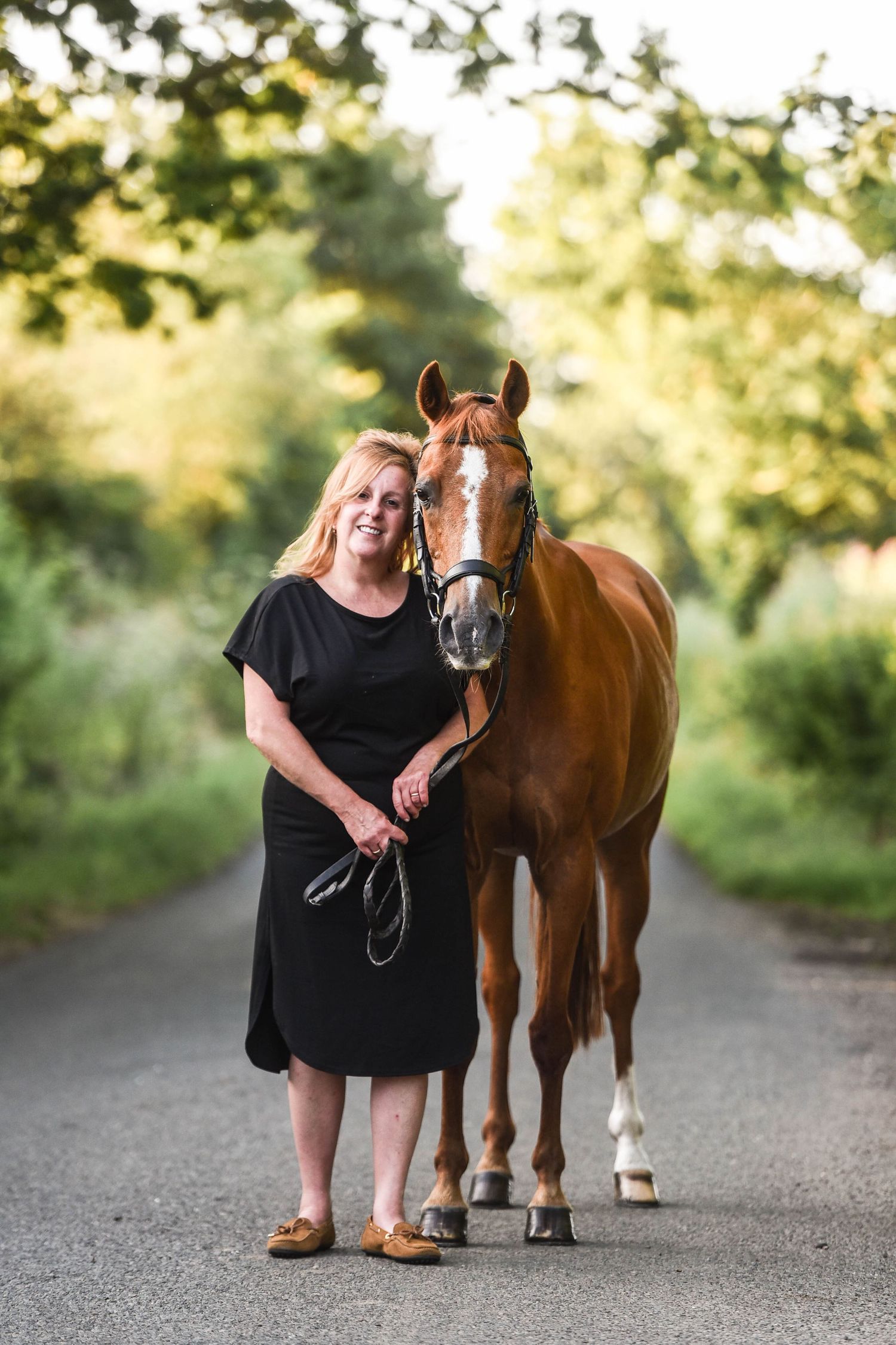 Smiling blonde woman in black dress standing beside a chestnut horse on a tree-lined country road.