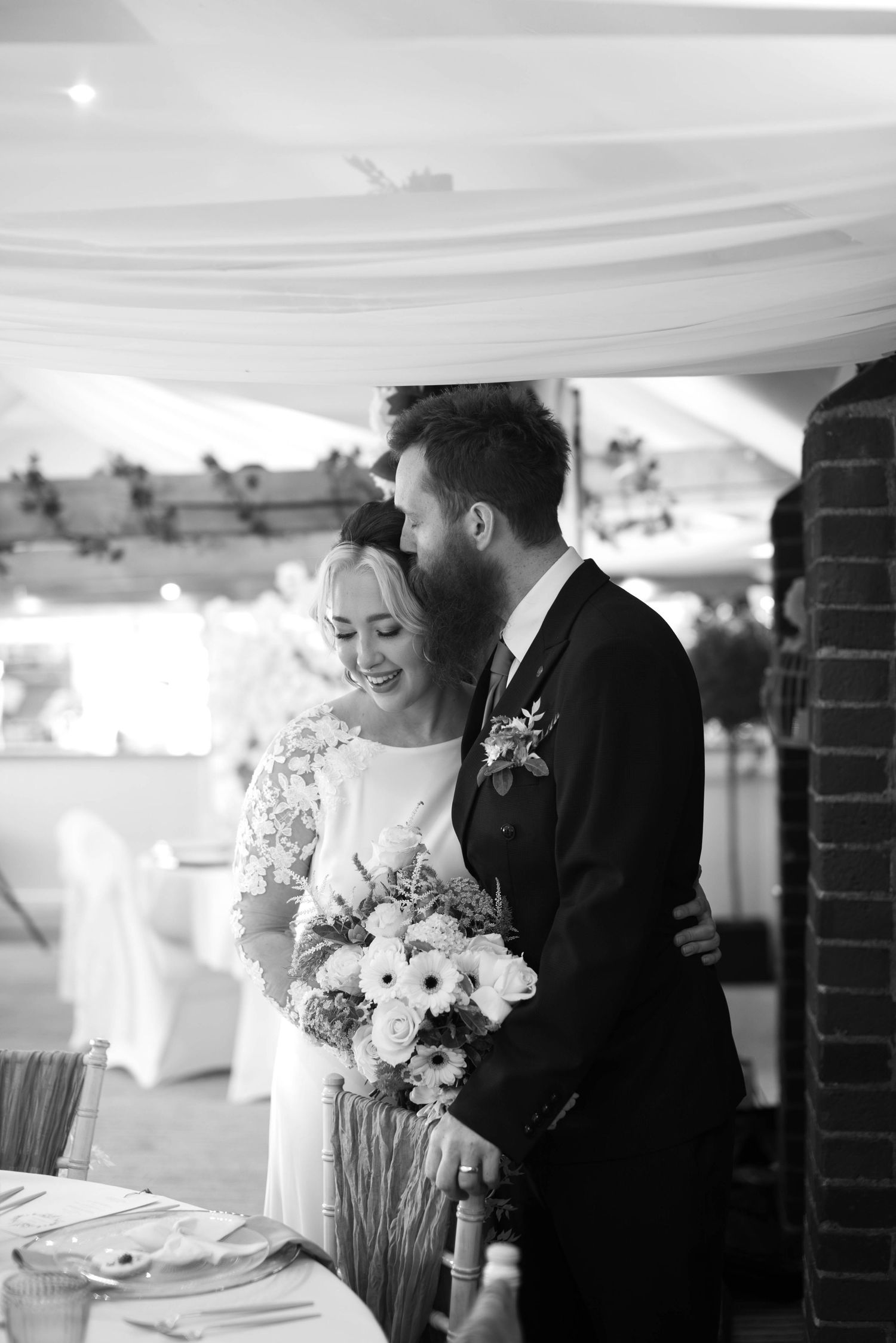 Black and white wedding photo of bride and groom sharing intimate moment beside reception table with bouquet.