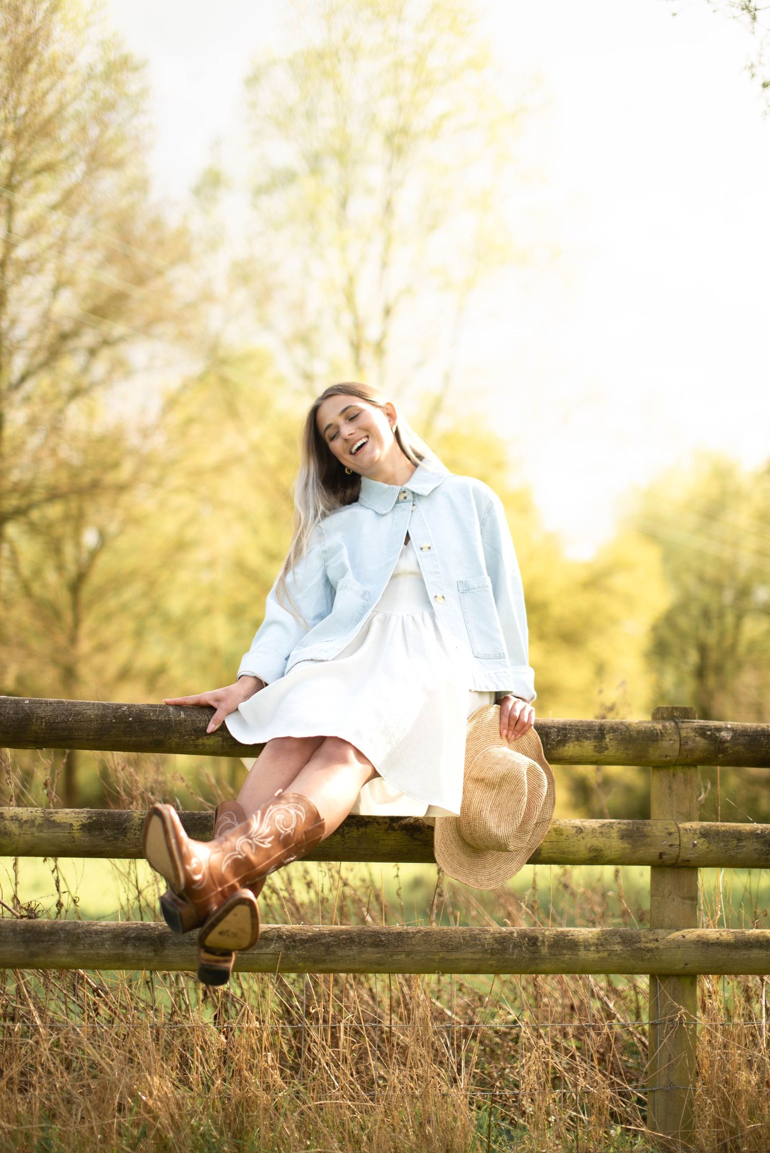 Smiling woman in white dress and denim jacket sits on wooden fence holding hat in sunny field.