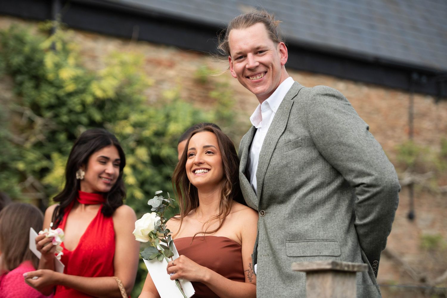 Smiling man in grey suit posing with a young woman holding white flowers at an outdoor wedding celebration.