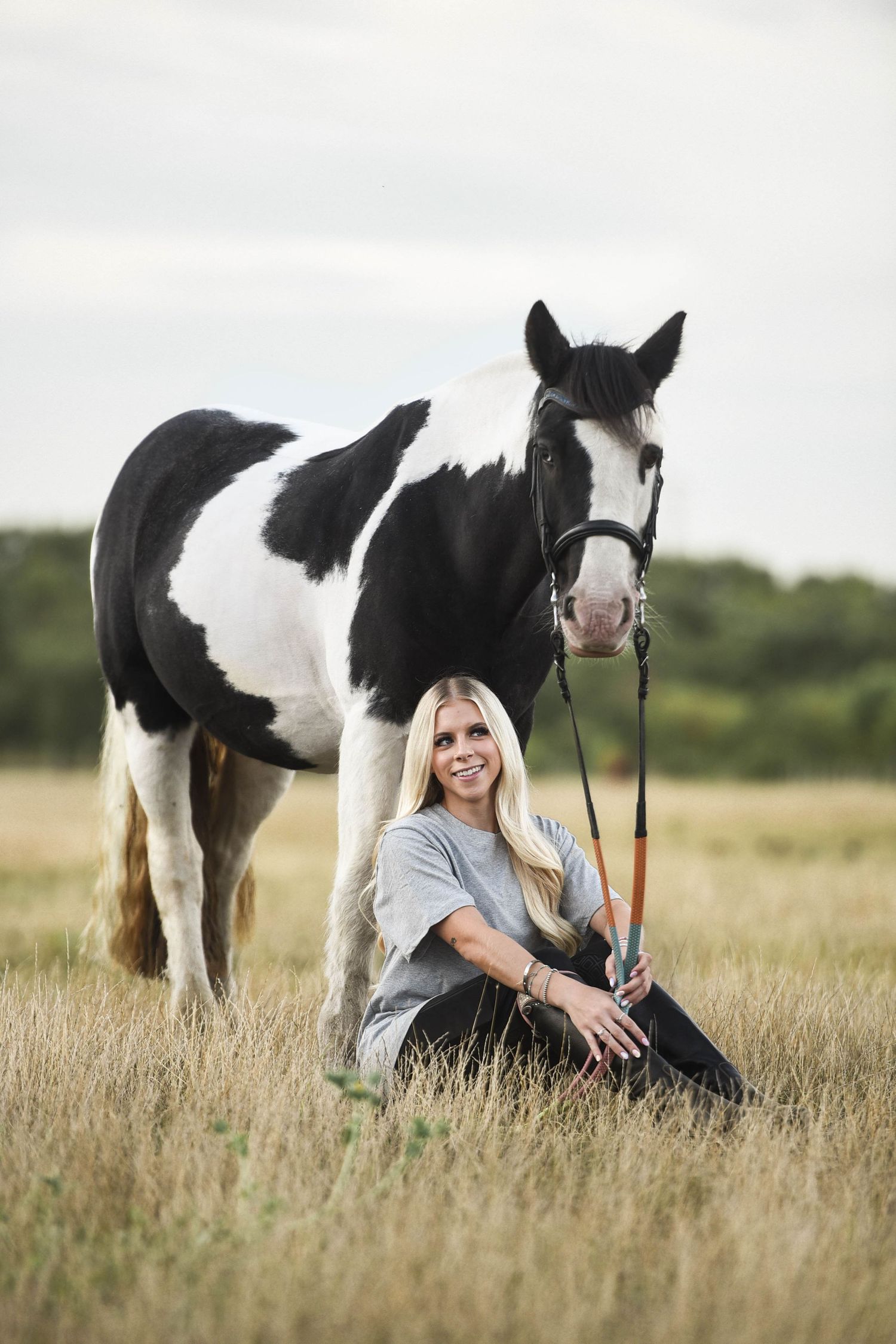 Smiling blonde woman sitting in field holding lead rope of black and white horse.