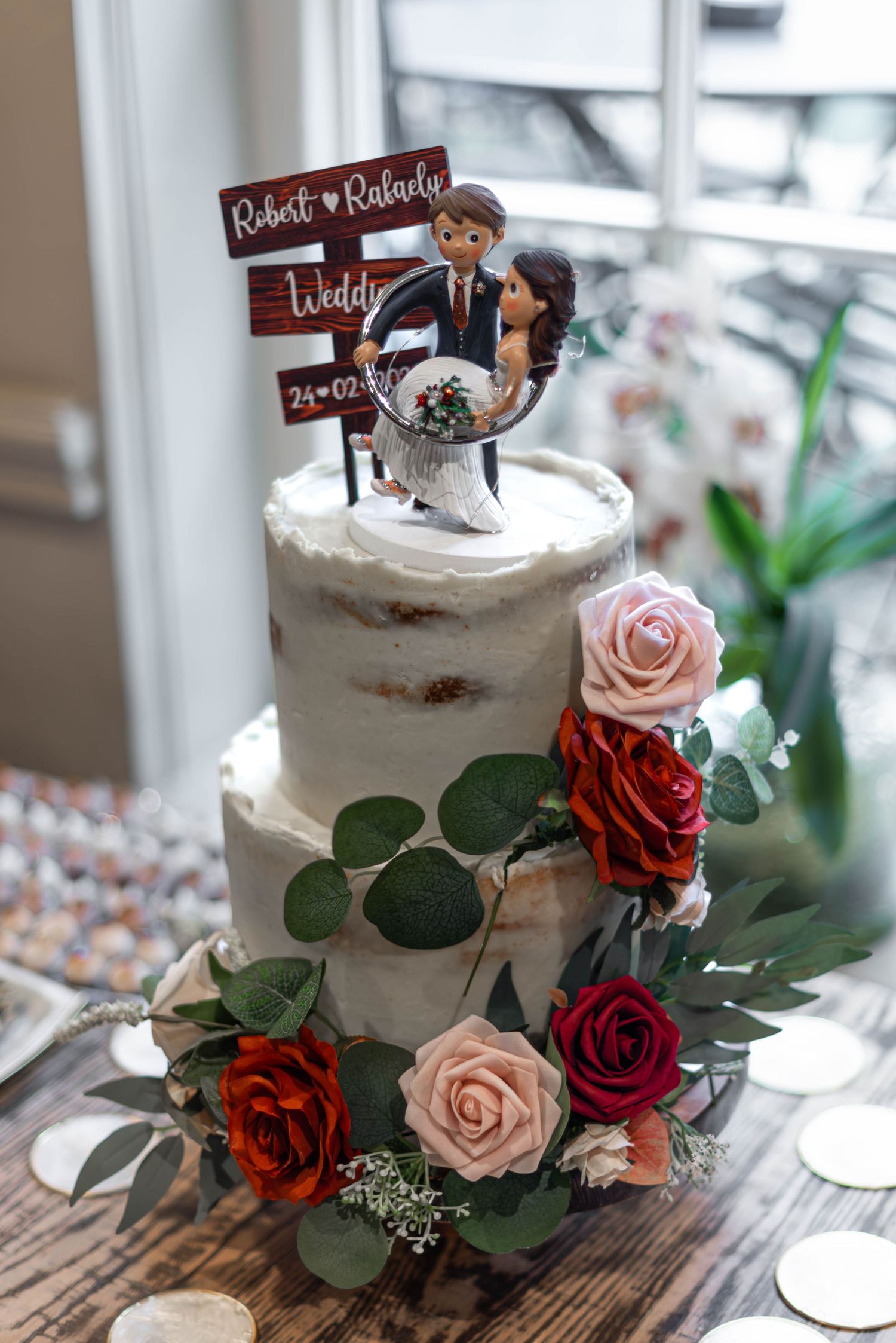 Two-tier naked wedding cake decorated with red and pink roses and custom cartoon cake topper.