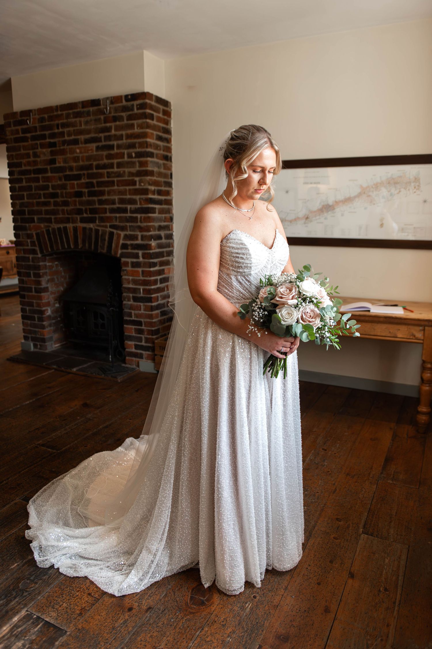 Bride in a strapless glitter ball gown holding a blush bouquet, standing by a brick fireplace on hardwood floors.