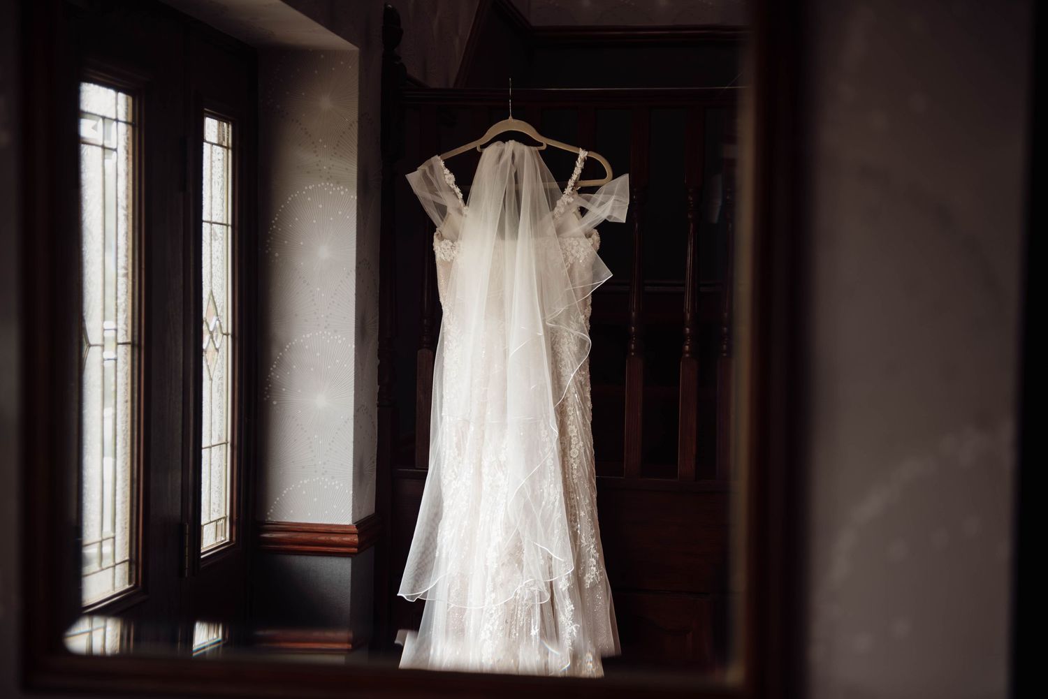 White lace wedding dress with veil hanging on a hanger in a dimly lit hallway near elegant windows.