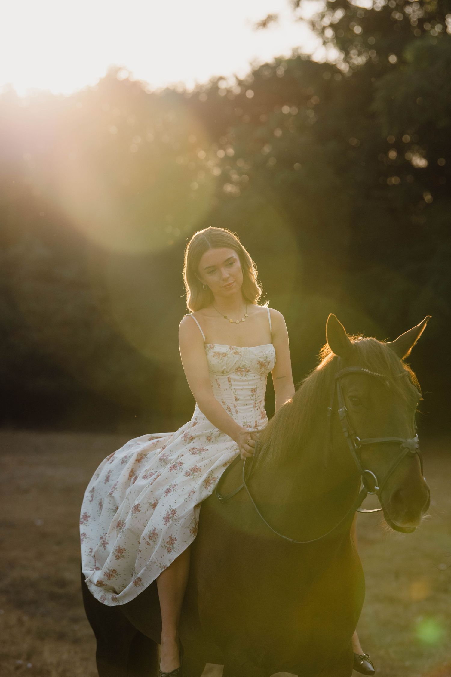 Young woman in floral dress riding a dark horse during golden hour sunset in a lush green field.