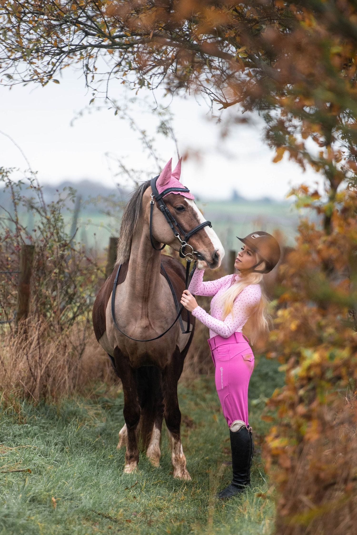A woman in pink equestrian attire and helmet stands beside a brown horse in an autumn countryside setting.