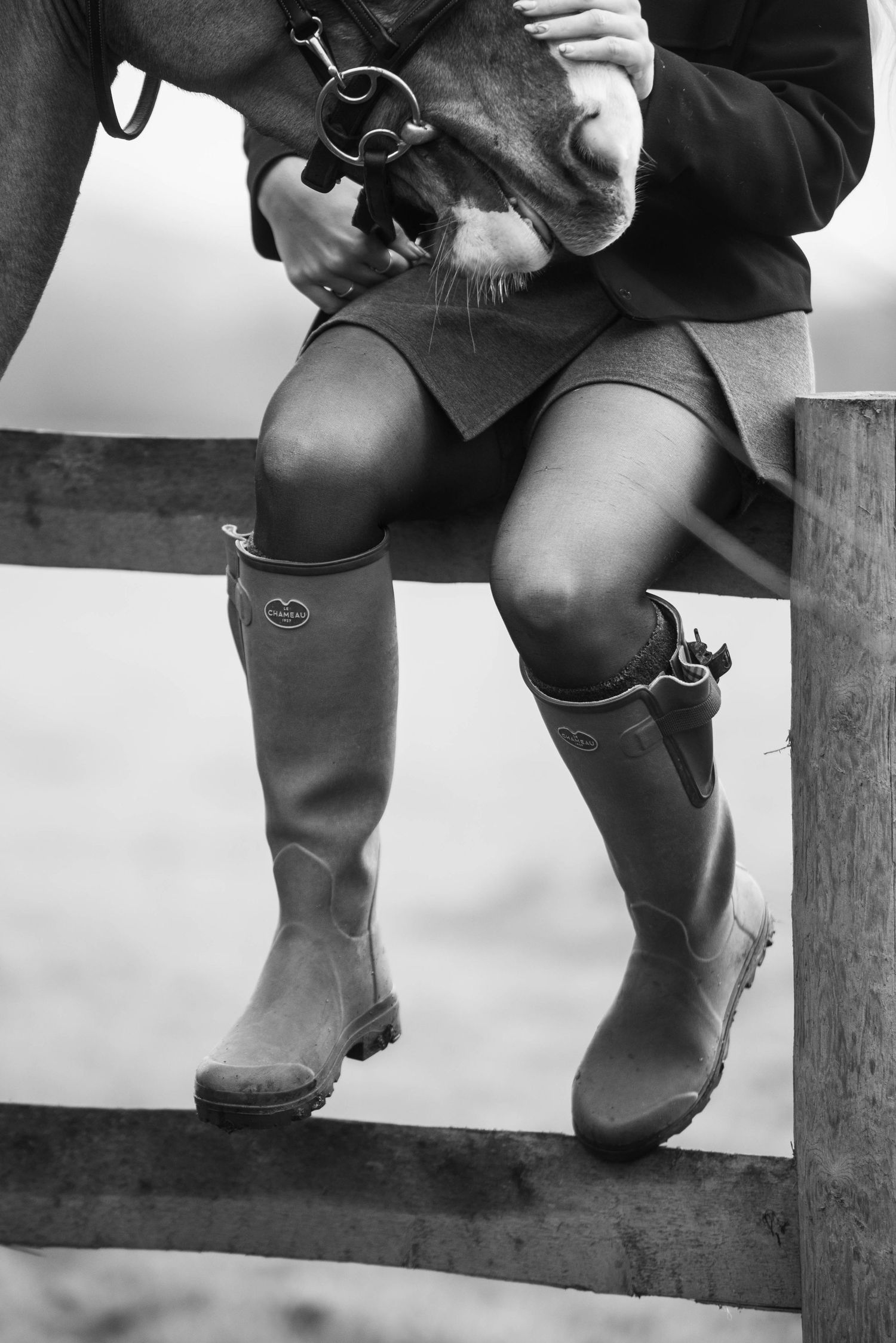 Black and white image of a person wearing tall rubber boots sitting on a wooden fence with a horse.