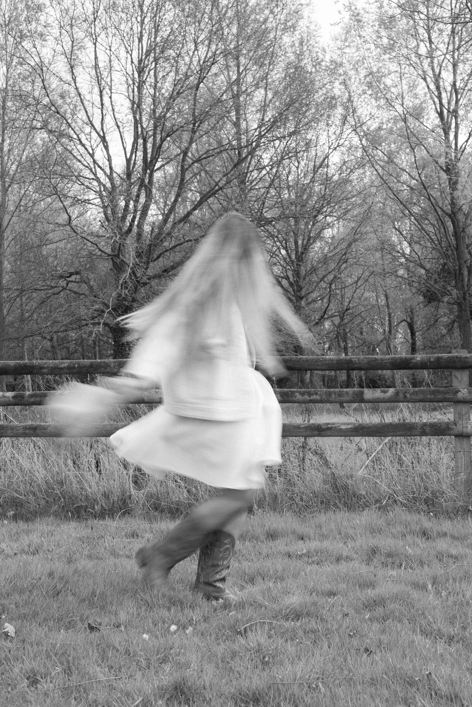 Motion blur black and white photo of woman in white dress spinning outdoors near wooden fence.