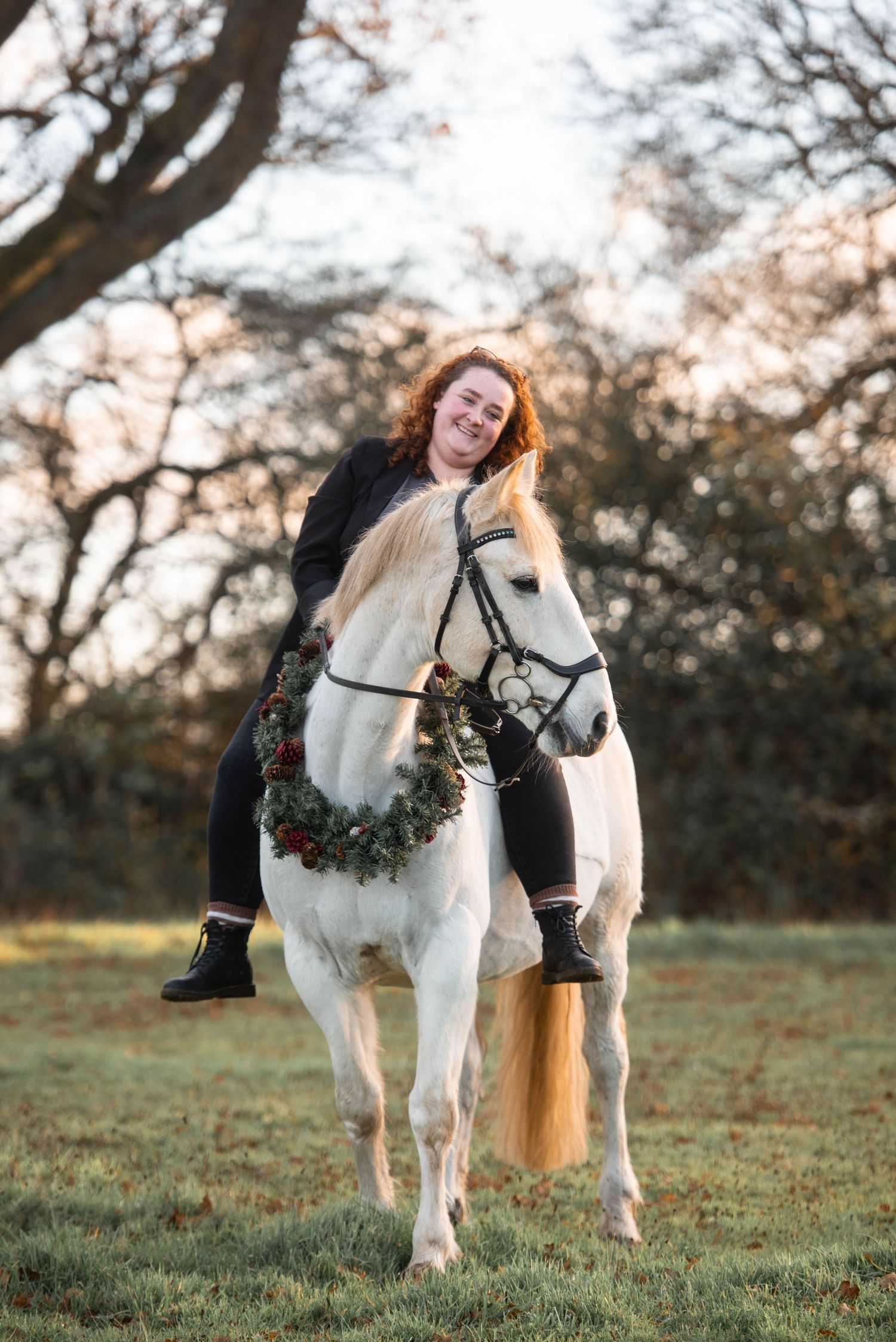 Smiling woman with curly red hair riding a white horse decorated with a Christmas wreath outdoors.