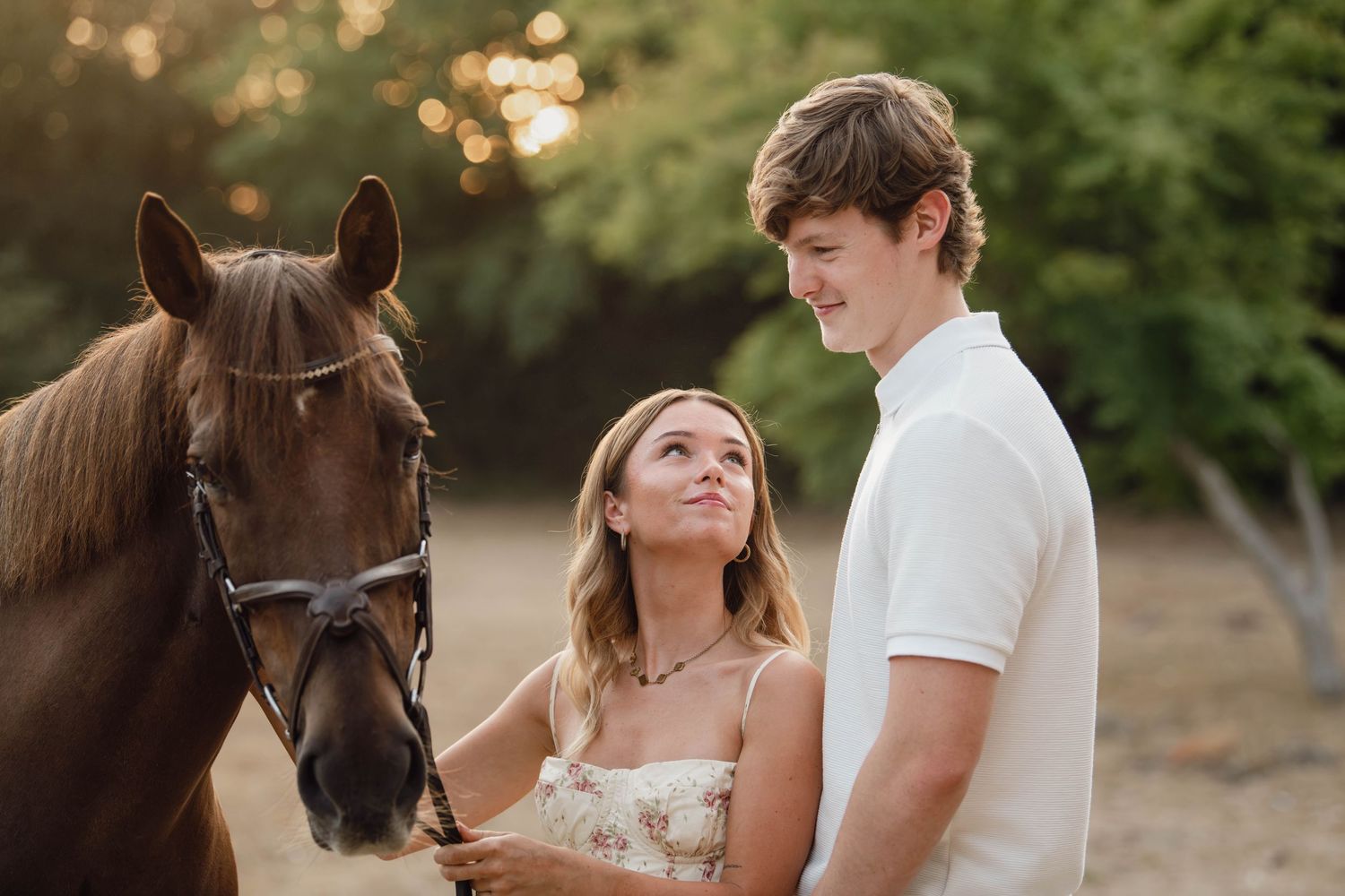 Young couple smiling together beside a brown horse outdoors in golden hour sunlight.