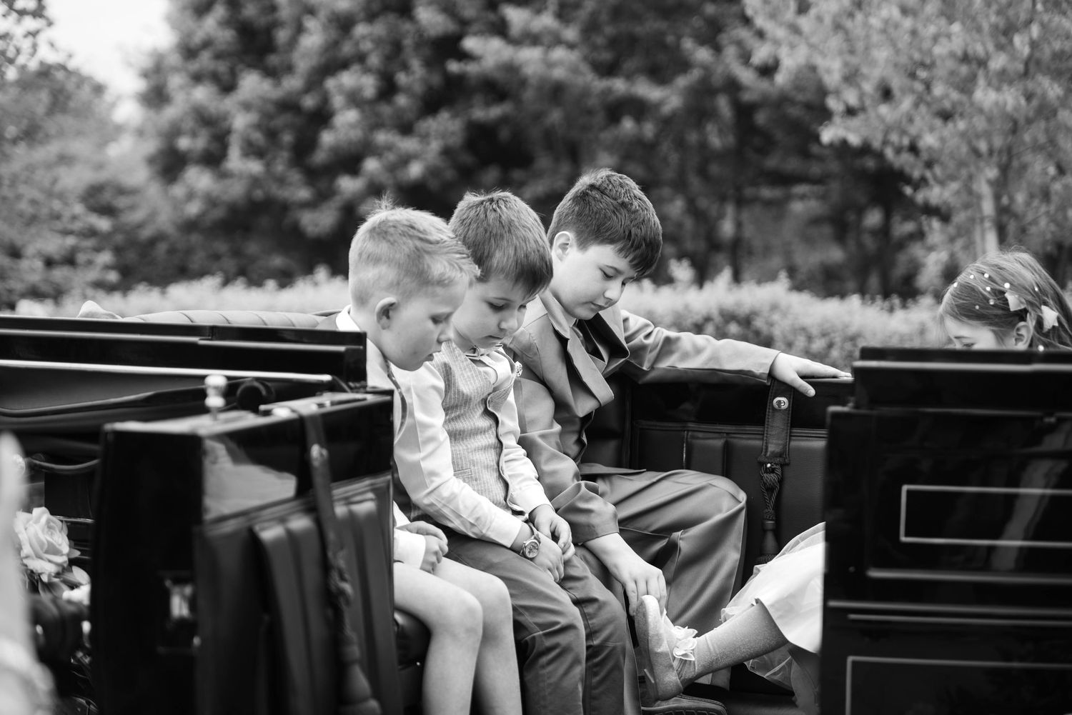 Three young boys in formal attire sit together on a bench at an outdoor wedding ceremony.