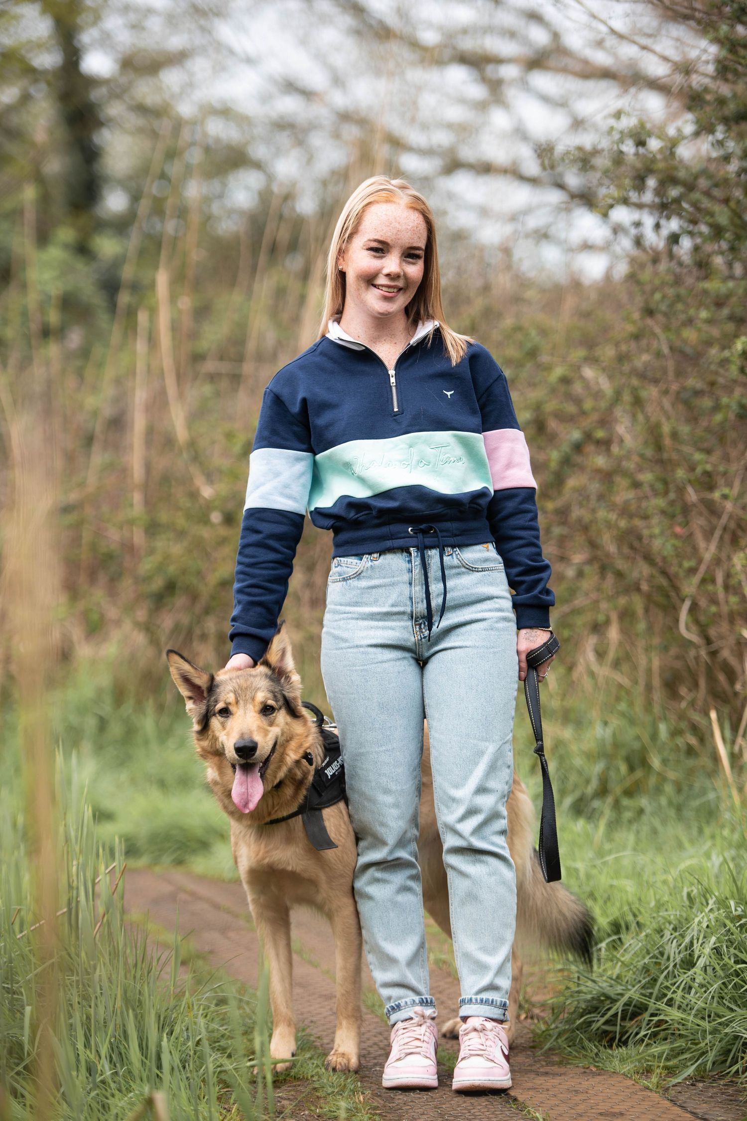 Young blonde woman in colorful sweatshirt walking a German Shepherd mix on a woodland path.