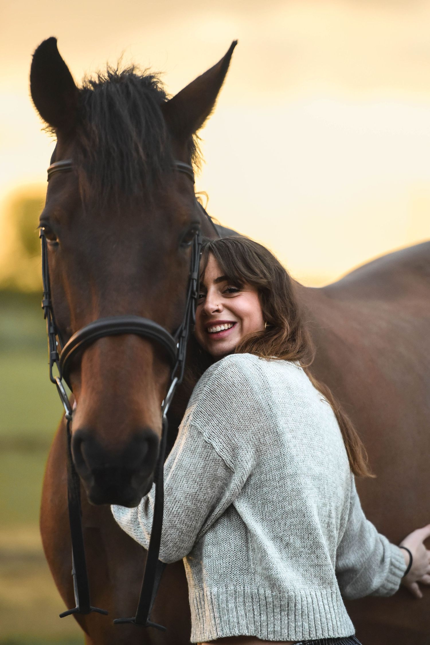 Smiling woman in grey sweater leans against a brown horse at sunset in an open field.