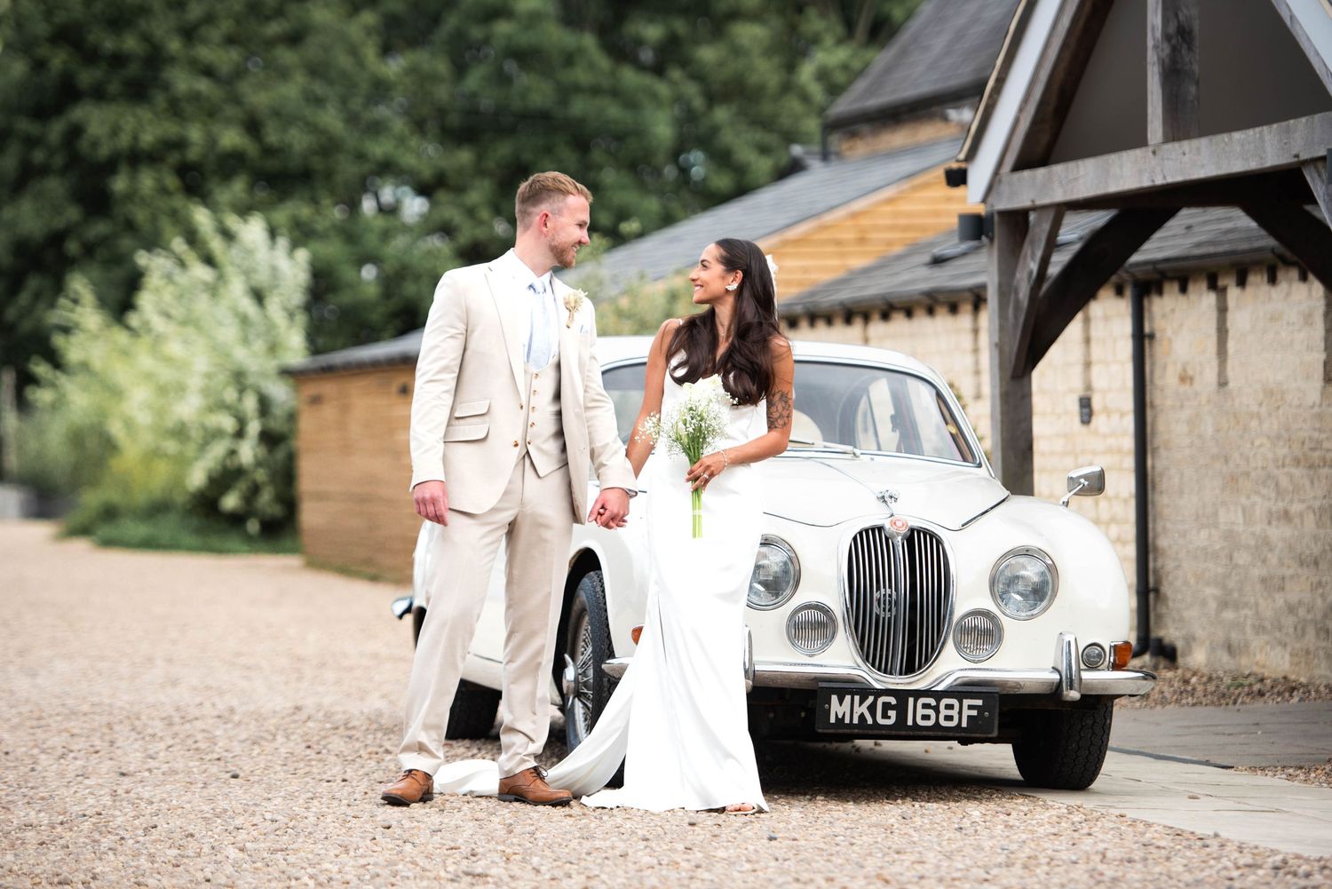 Bride and groom holding hands beside a vintage white Jaguar classic car on a gravel driveway at a barn venue.