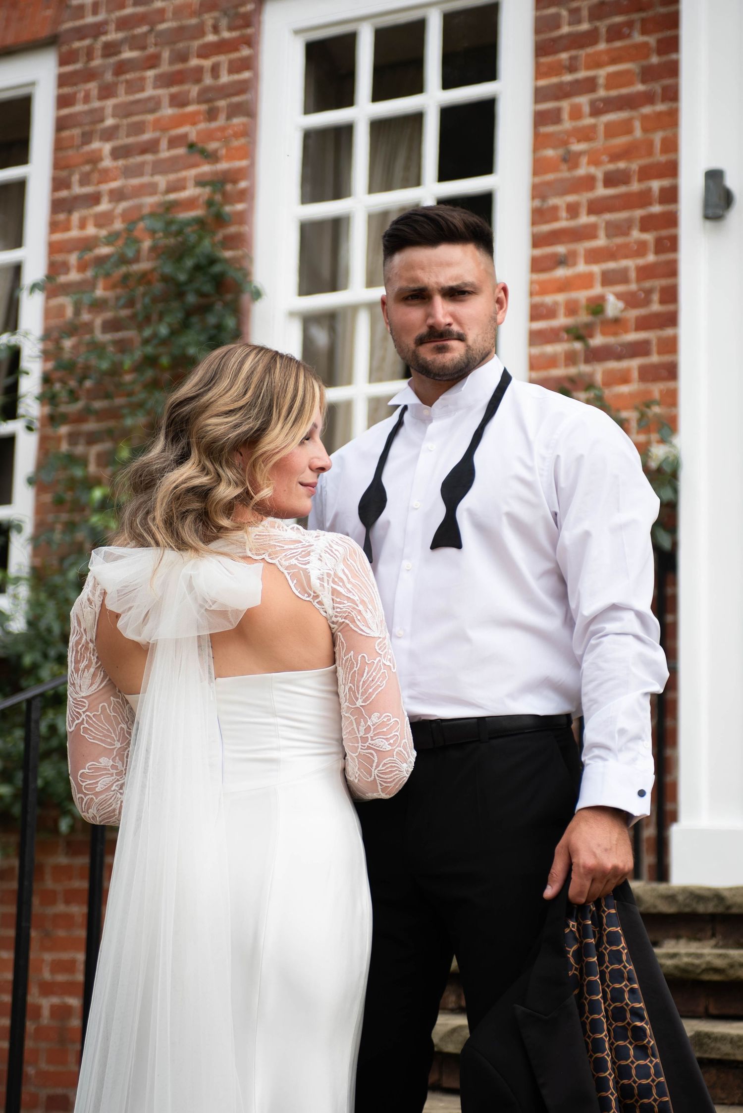 Groom in white shirt and black suspenders stands with bride in lace wedding dress outside brick building.