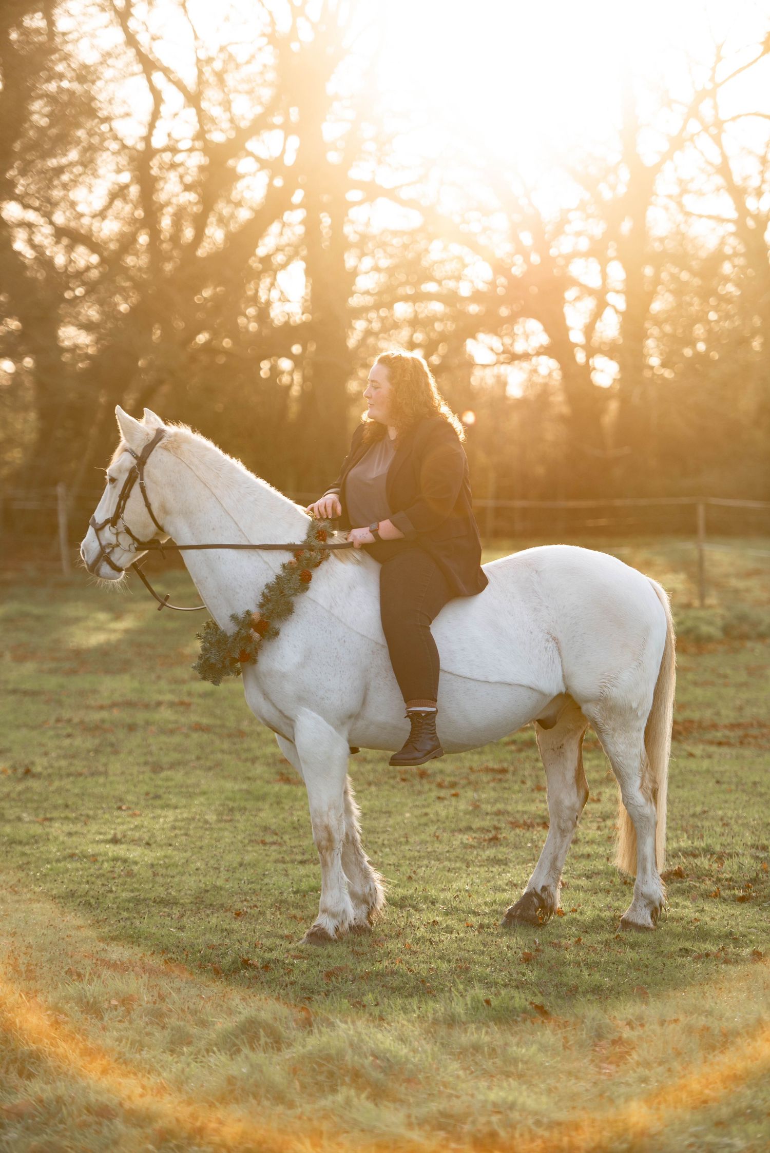 Woman in black riding a white horse adorned with a festive wreath at golden hour sunset.