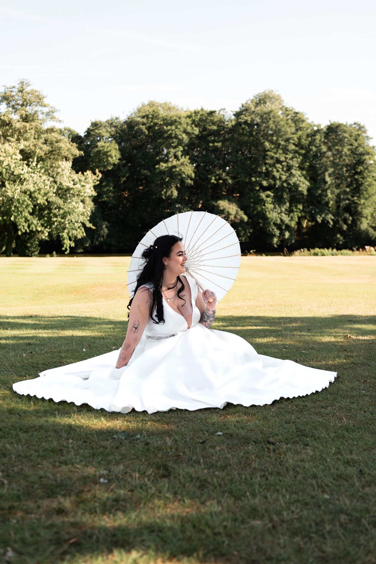 Bride in white gown sits on grass holding white parasol umbrella, surrounded by lush green trees on a sunny day.