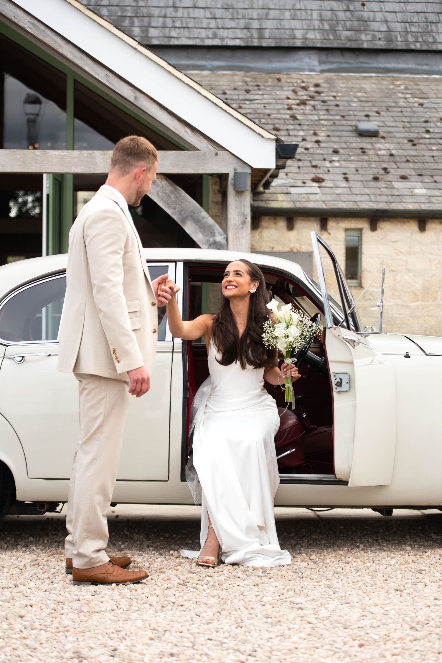 A bride in a white gown holds a bouquet while stepping out of a vintage car assisted by the groom.