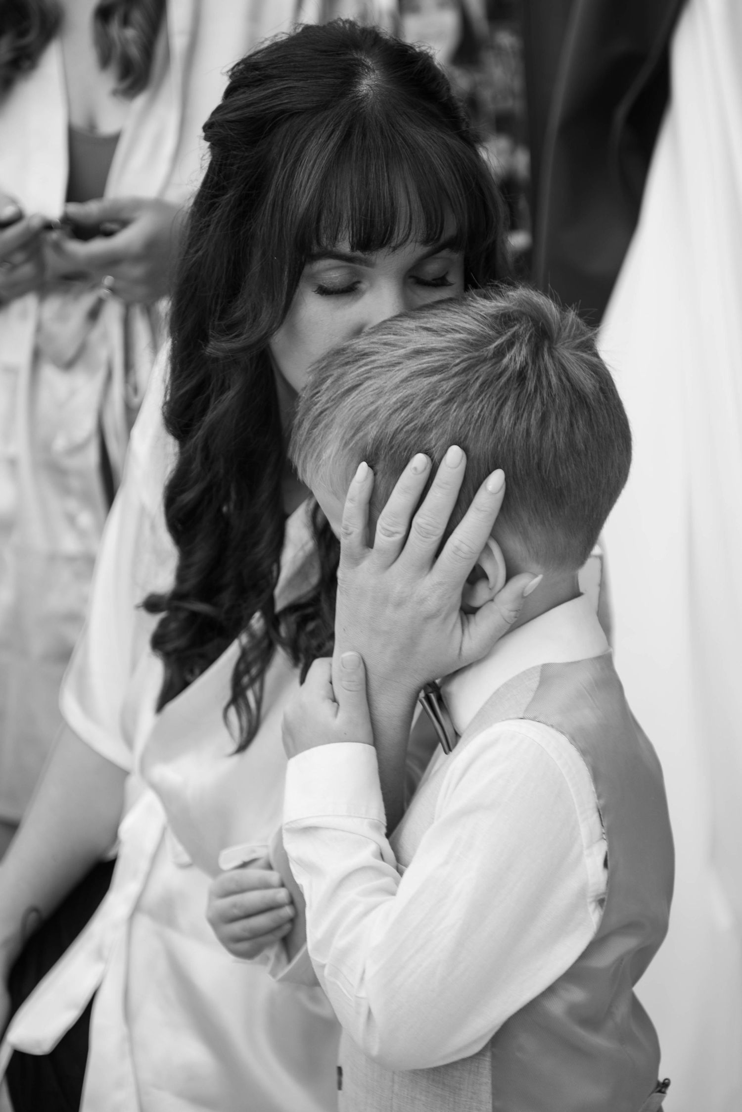 Black and white photo of dark-haired woman in satin robe tenderly embracing young boy at wedding.
