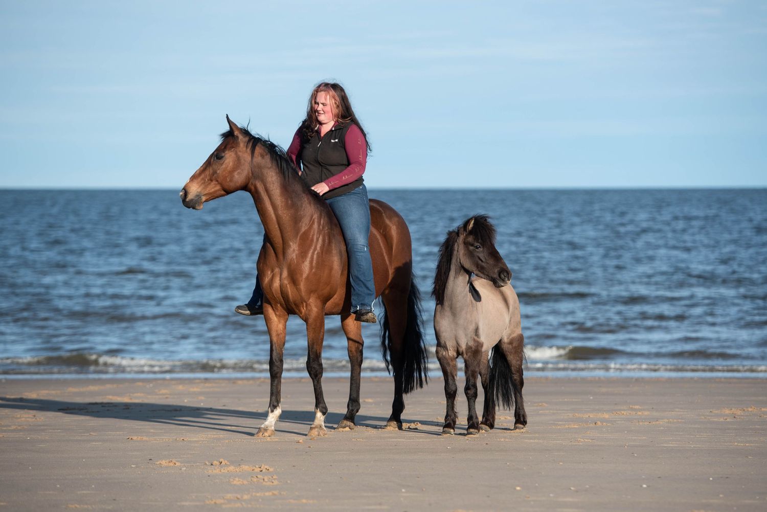 Woman riding a bay horse on a sandy beach with a small dark pony walking alongside.