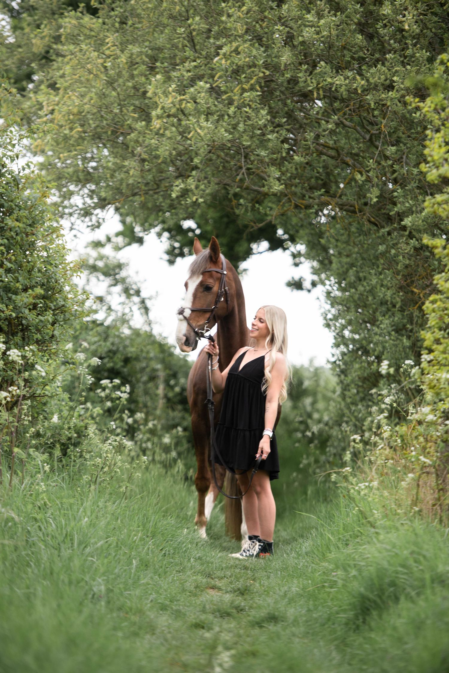 Blonde woman in black dress standing beside a chestnut horse on a green leafy trail.
