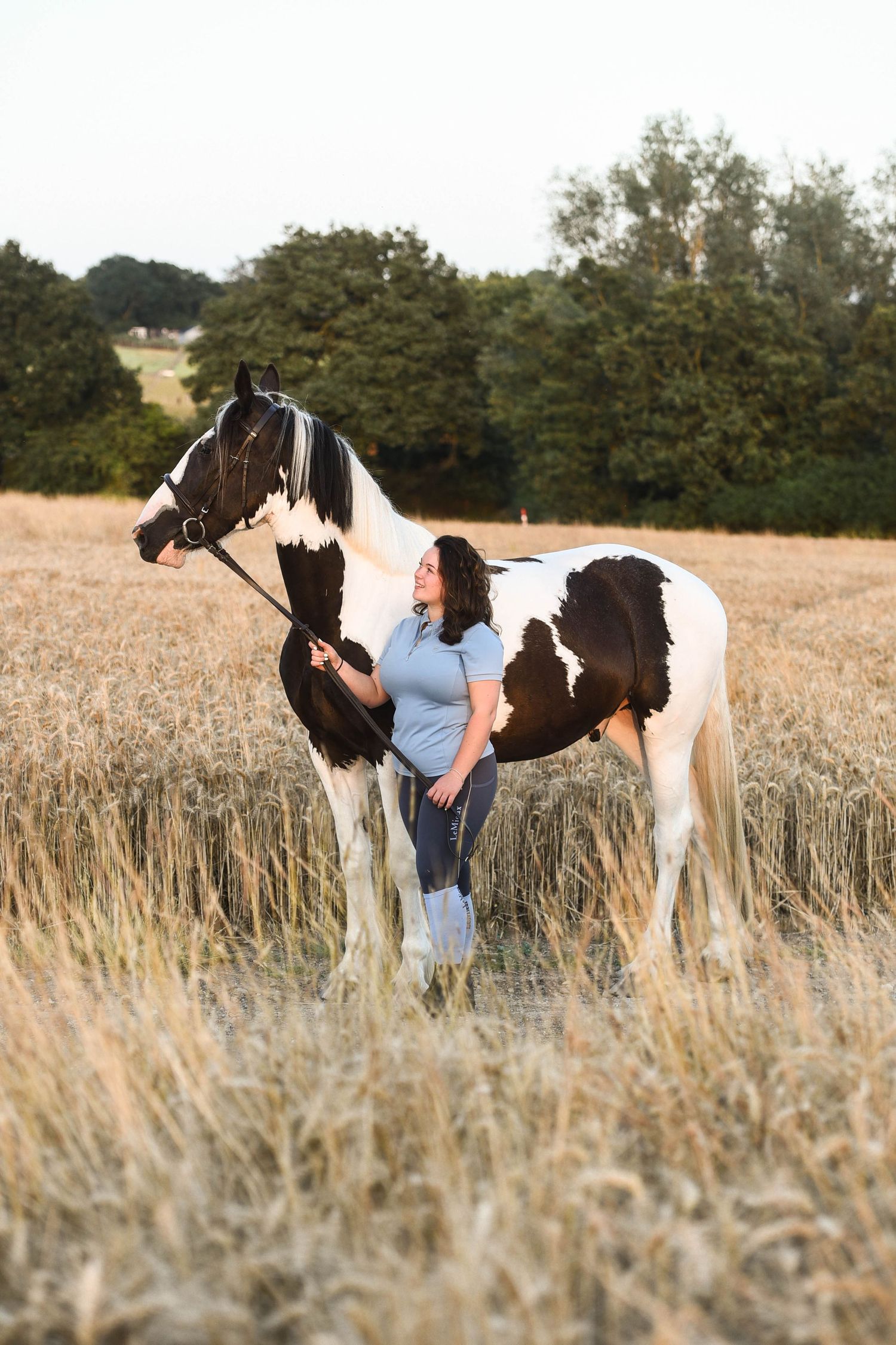 A woman stands beside a black and white paint horse in a harvested golden wheat field.
