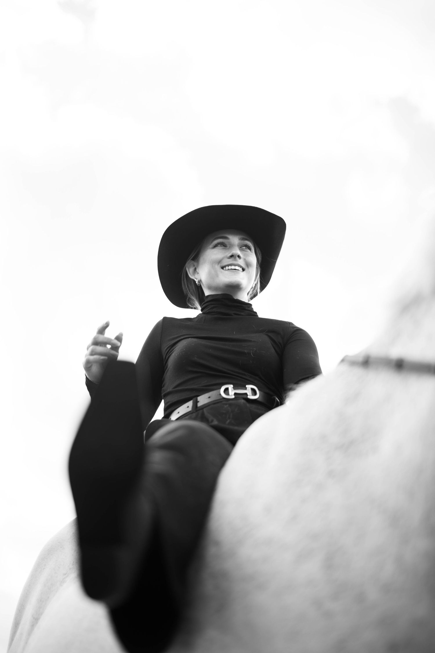 Low angle black and white photo of smiling woman in black cowboy hat riding a white horse.