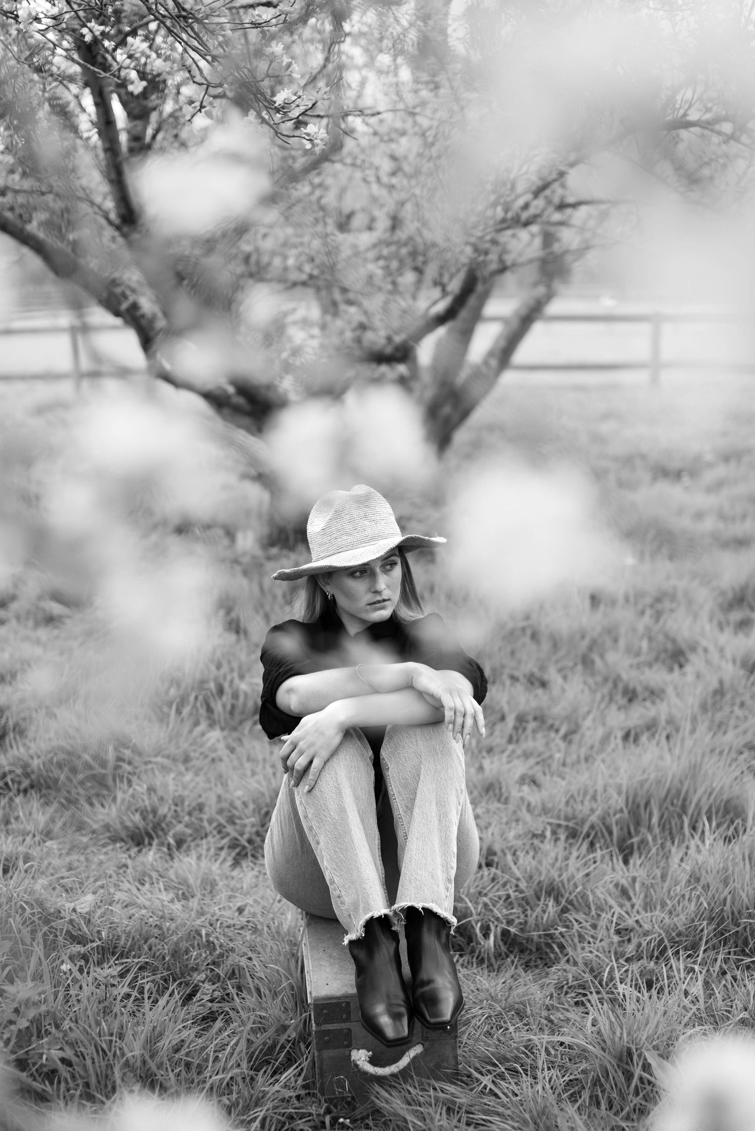 Black and white portrait of woman in straw hat and boots sitting cross-legged in grassy orchard.