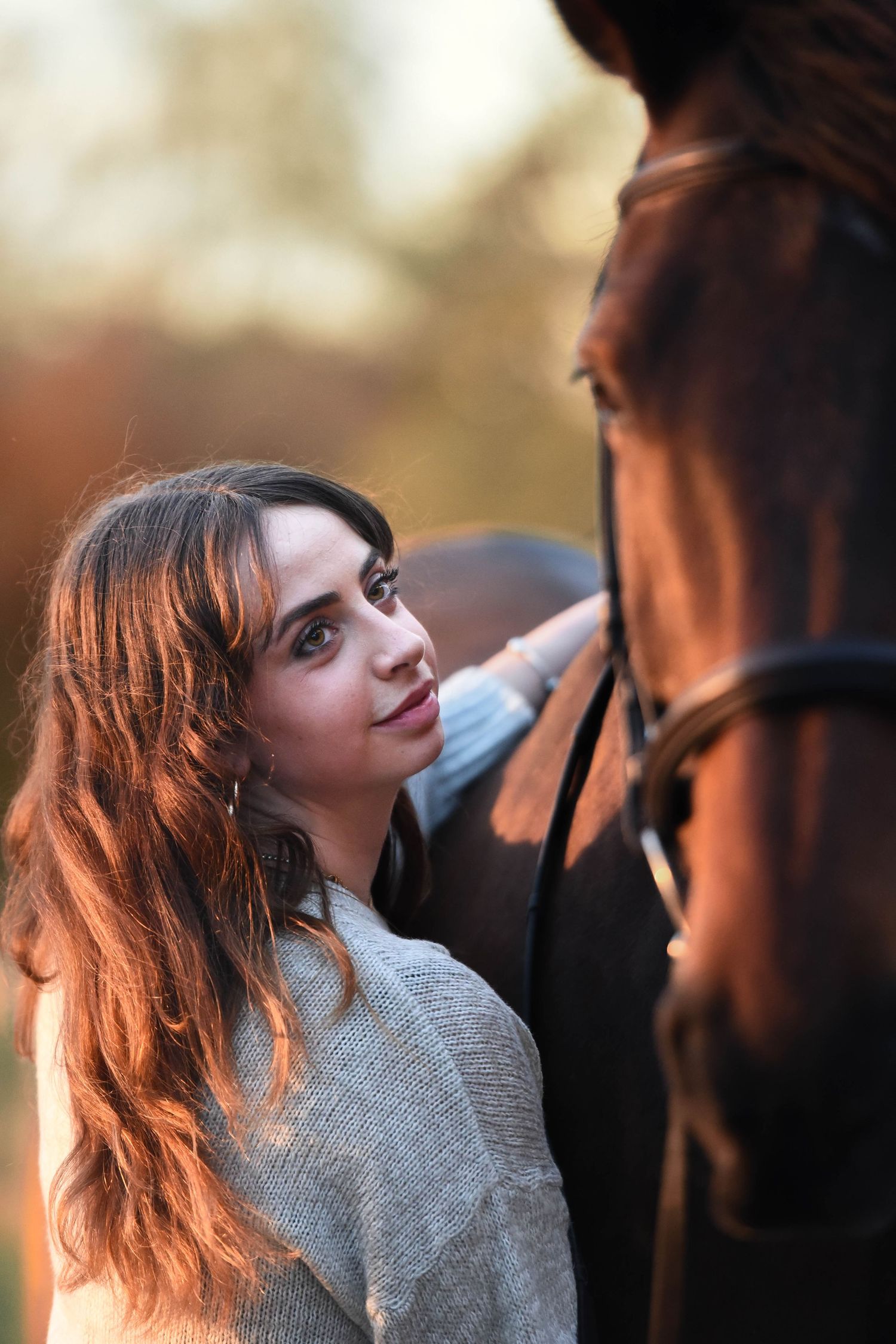 Young woman with long hair gently touches a brown horse's face during golden hour outdoors.