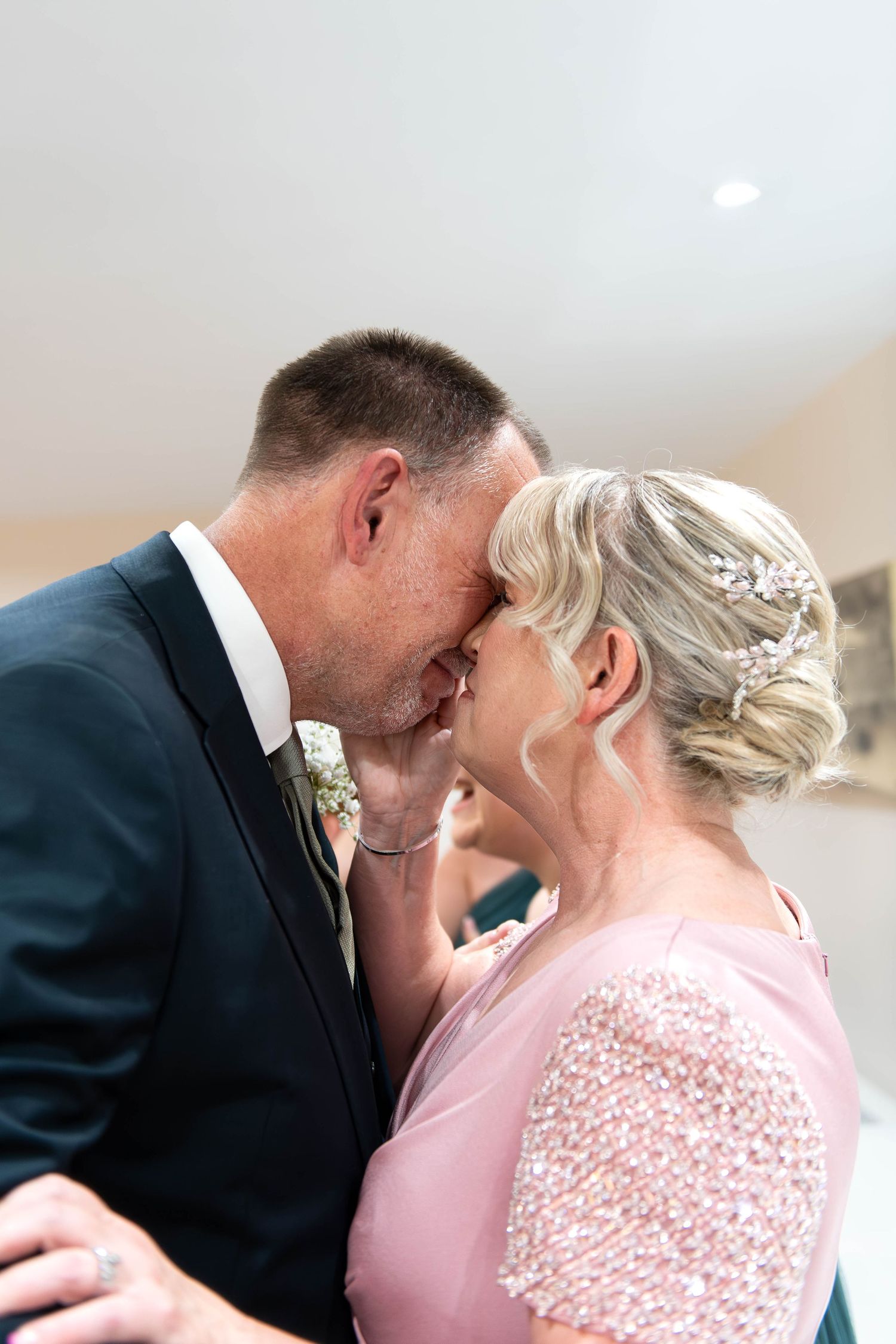 Couple sharing a romantic kiss at a wedding, groom in navy suit with boutonniere, bride in pink beaded dress with crystal hair piece.