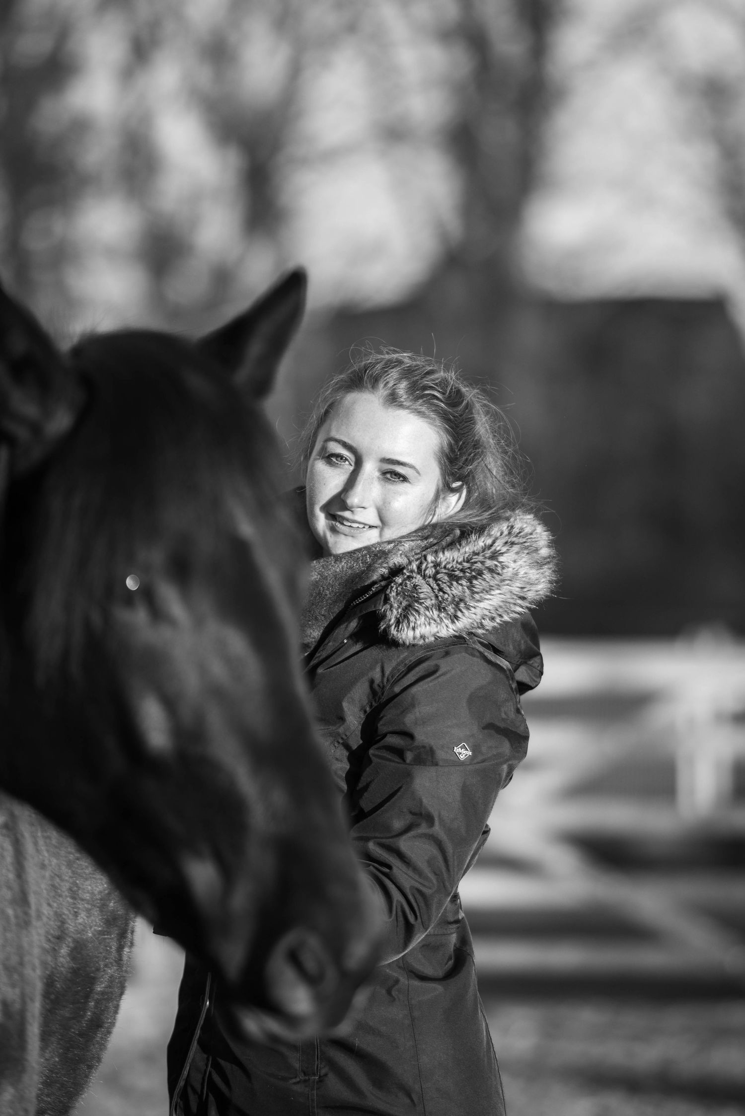 Smiling woman in winter jacket standing close to a dark horse in black and white photo.
