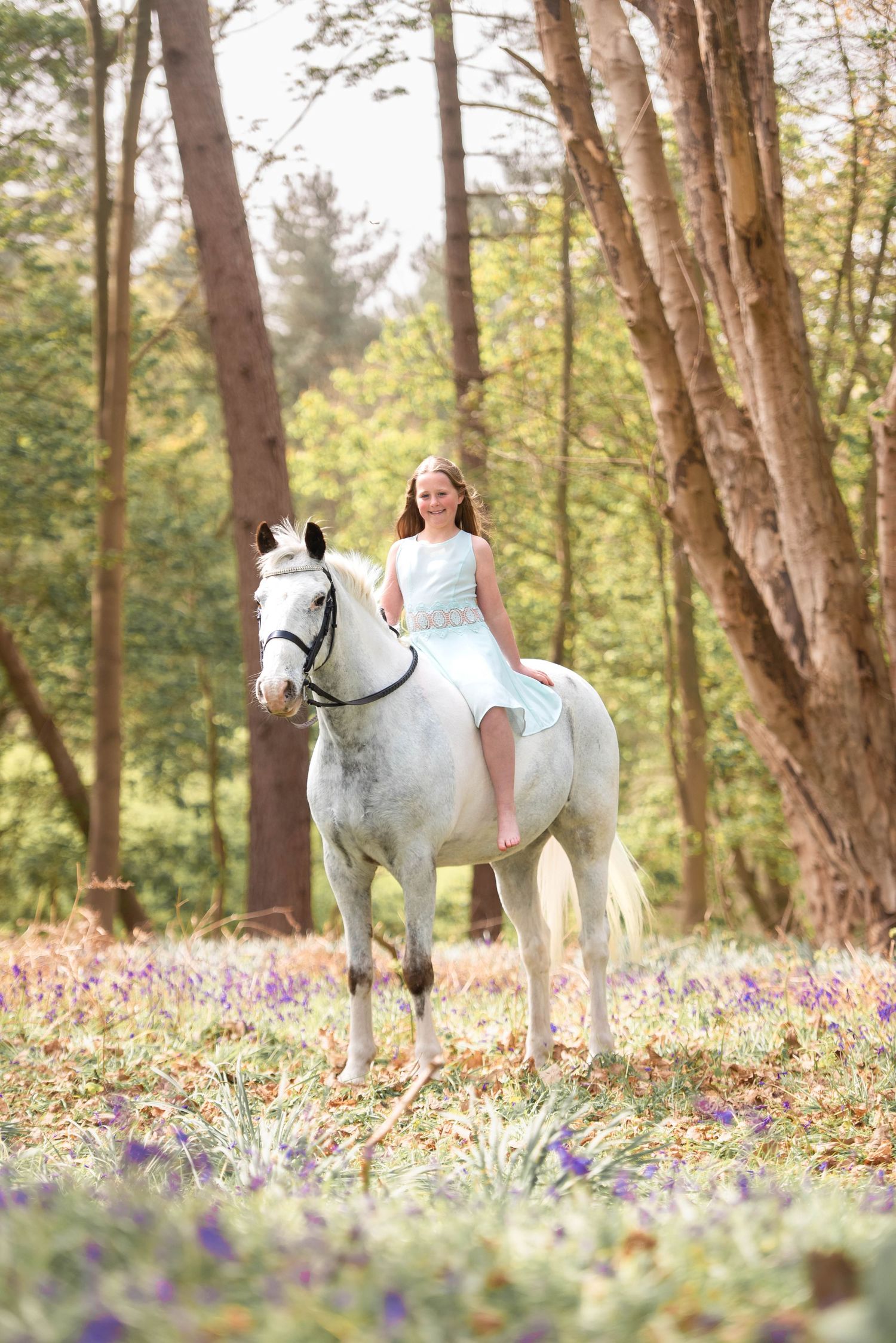 Smiling girl in blue dress riding a white horse through a bluebell woodland.