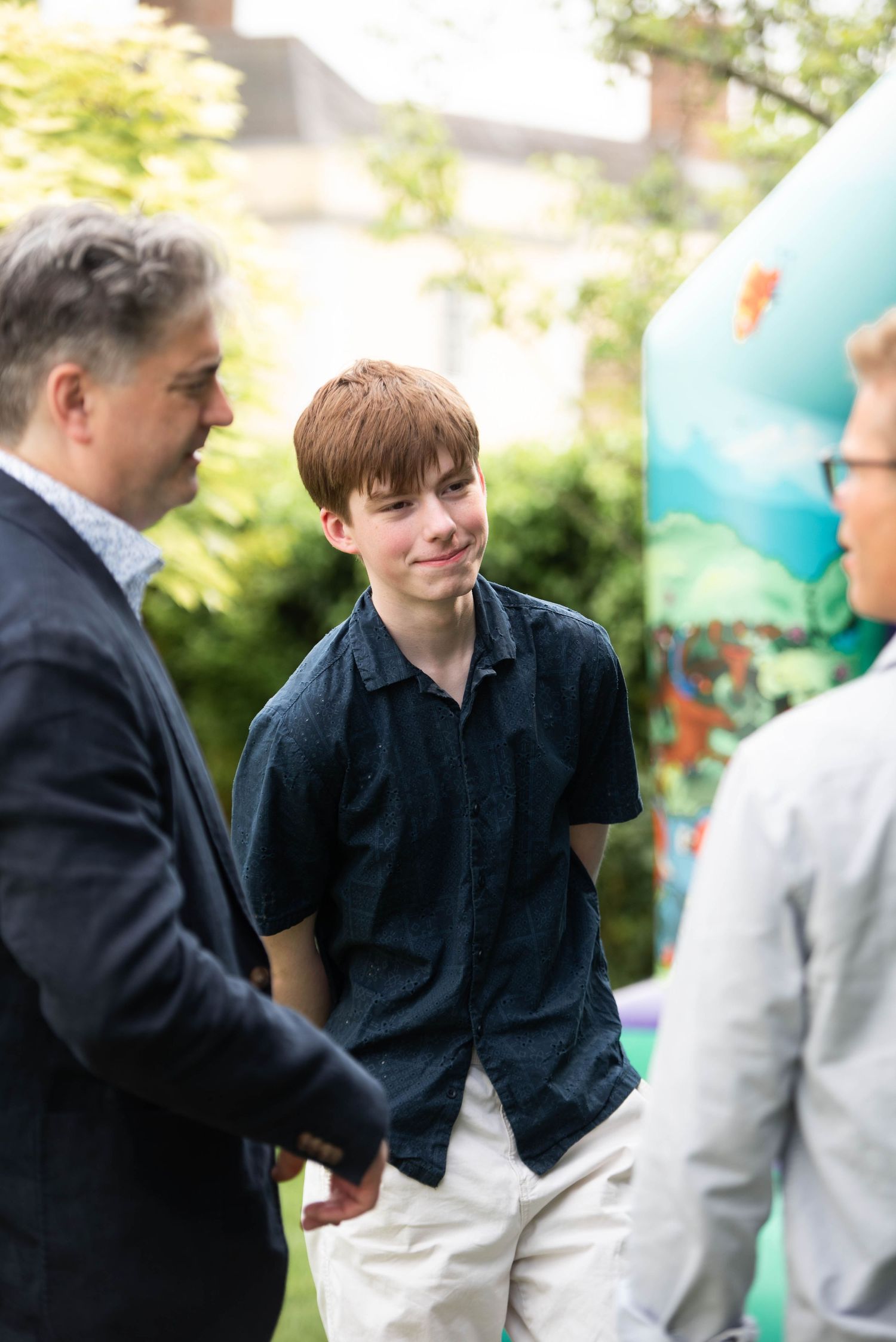 Teenage boy with red hair smiling while chatting with adults at an outdoor event.