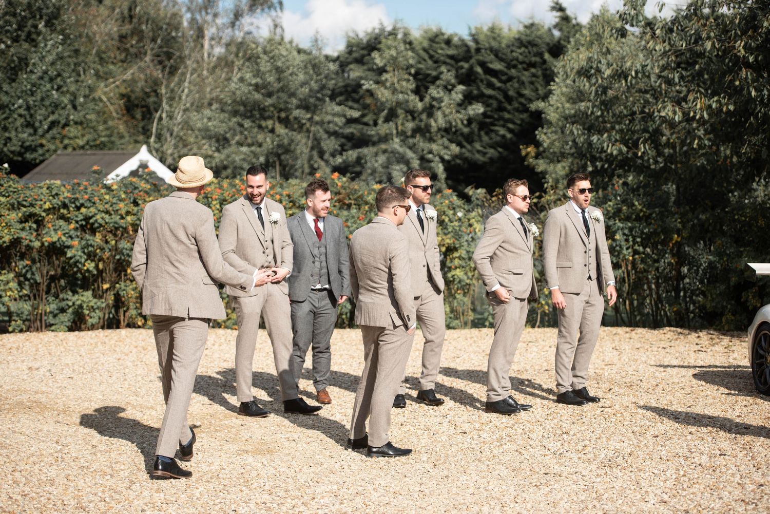 Groomsmen in matching tan suits socializing outdoors on gravel at a wedding venue.