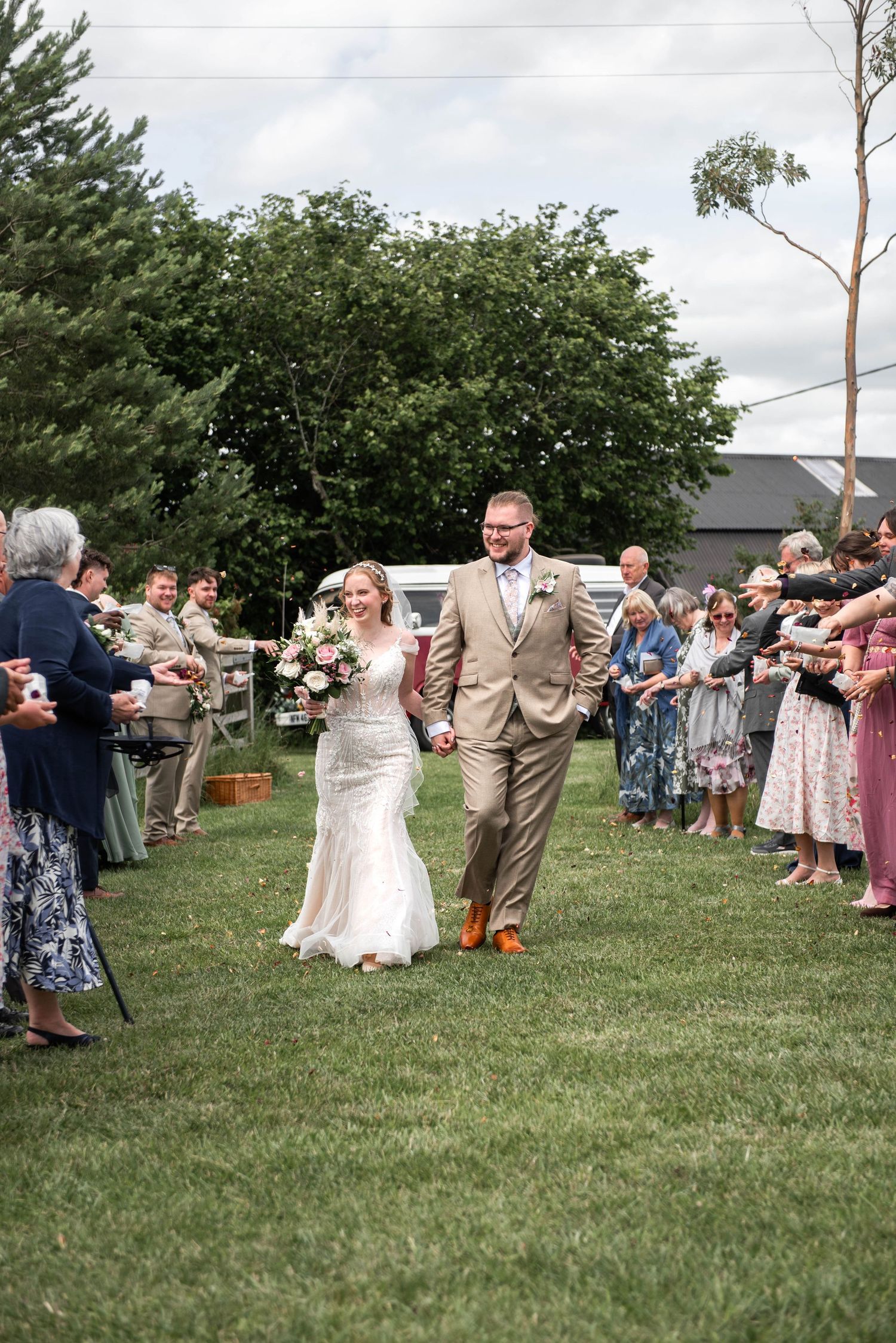 Bride and groom walk down outdoor aisle as guests celebrate their wedding ceremony on a sunny day.