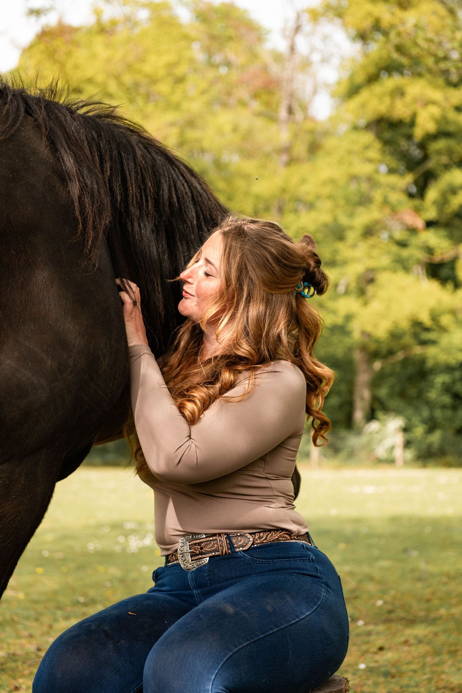 Woman in jeans hugging a black horse outdoors, smiling joyfully in a lush green park setting.
