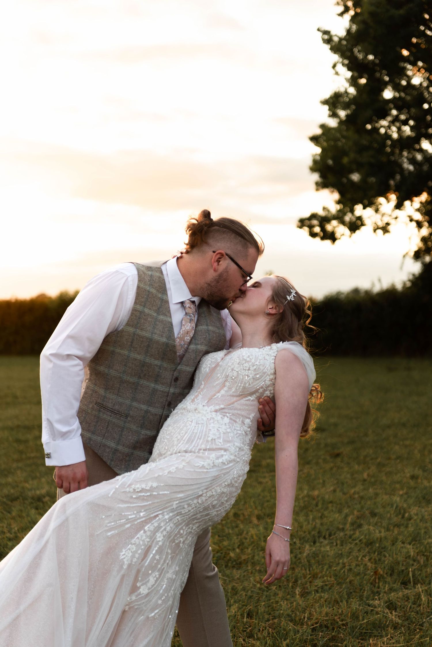 A groom dips his bride for a romantic kiss at sunset in a lush green field on their wedding day.
