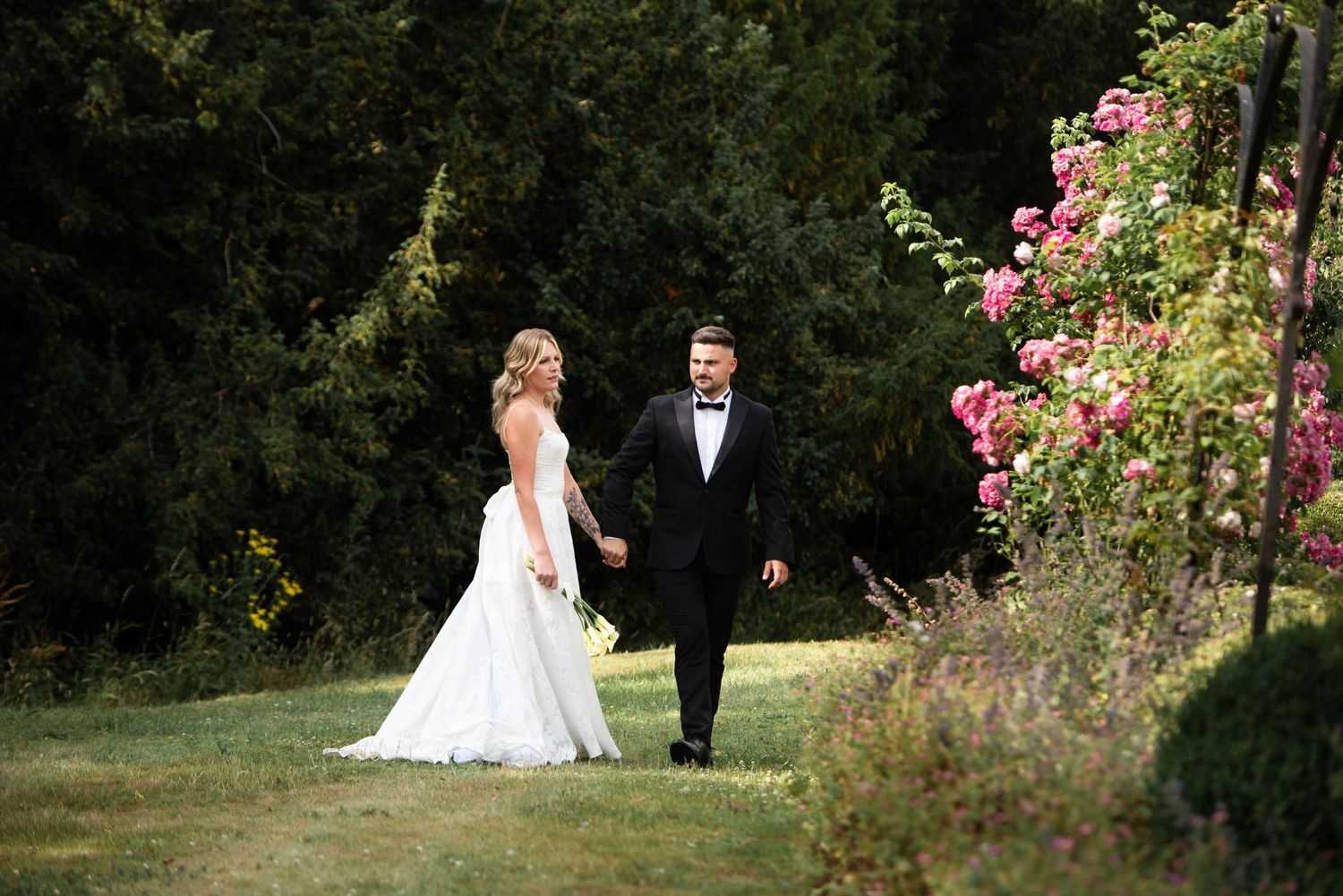 Bride and groom walk hand in hand through lush garden with blooming pink roses on their wedding day.
