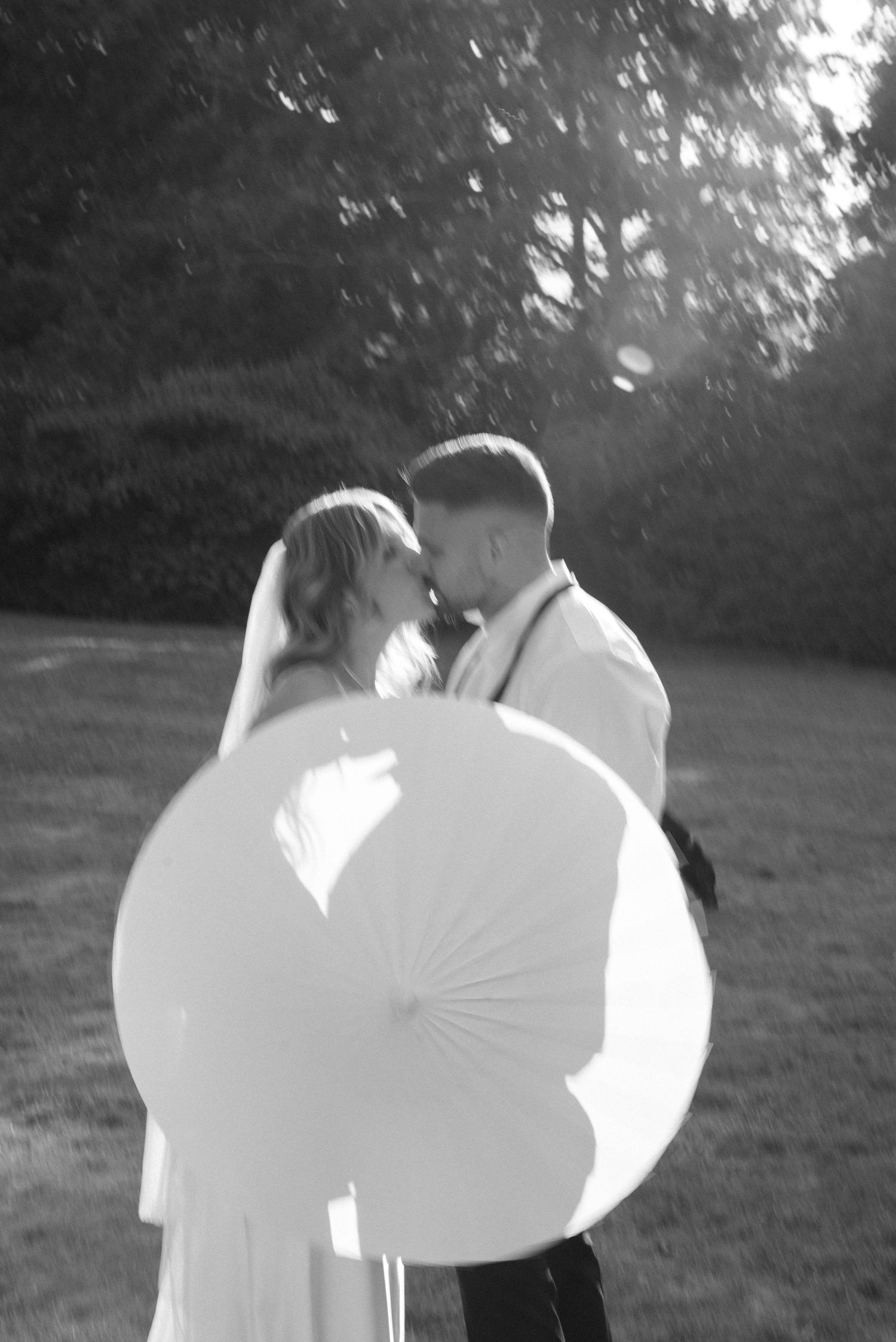 Black and white wedding photo of bride and groom with parasol in sunlit garden.